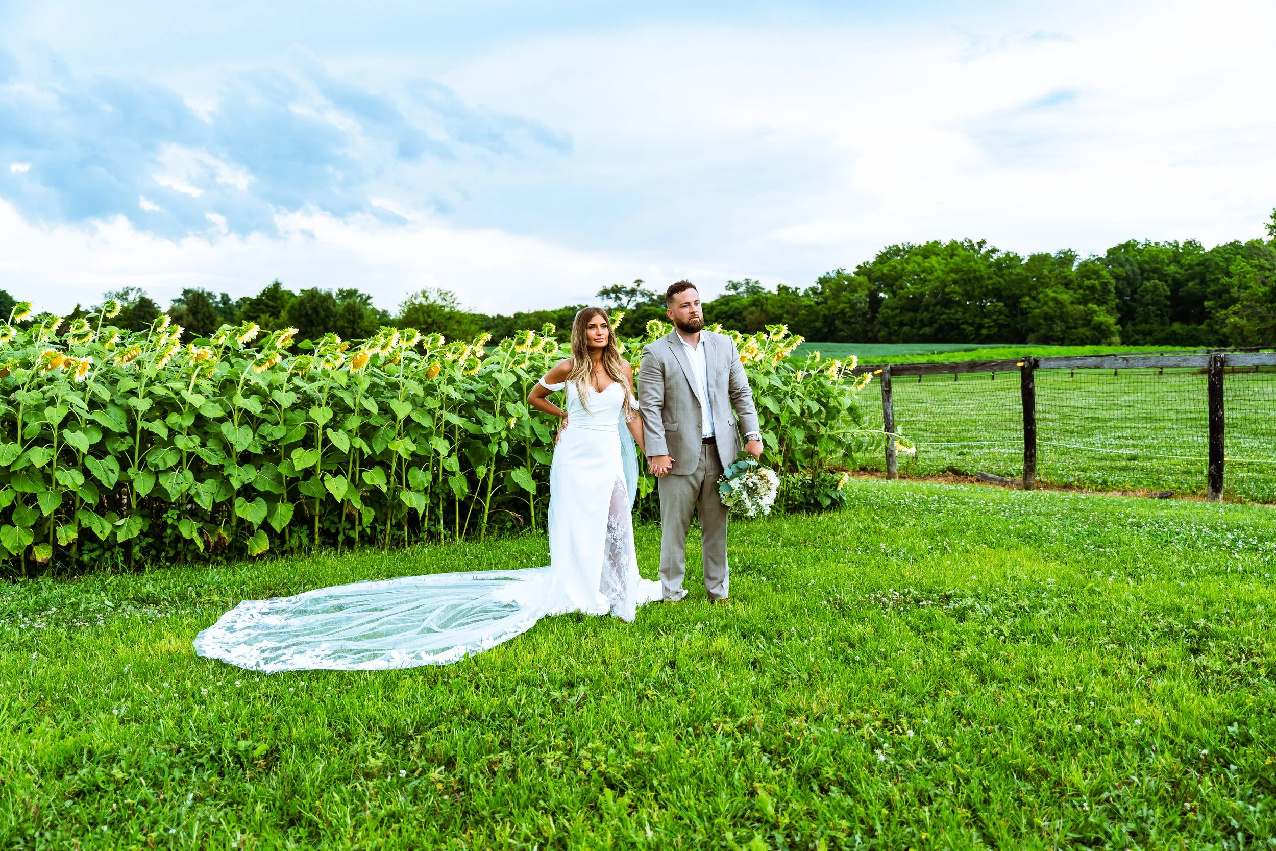 A bride and groom holding hands outdoors during a wedding, standing on grass near a sunflower field with green trees and a partly cloudy sky in the background.