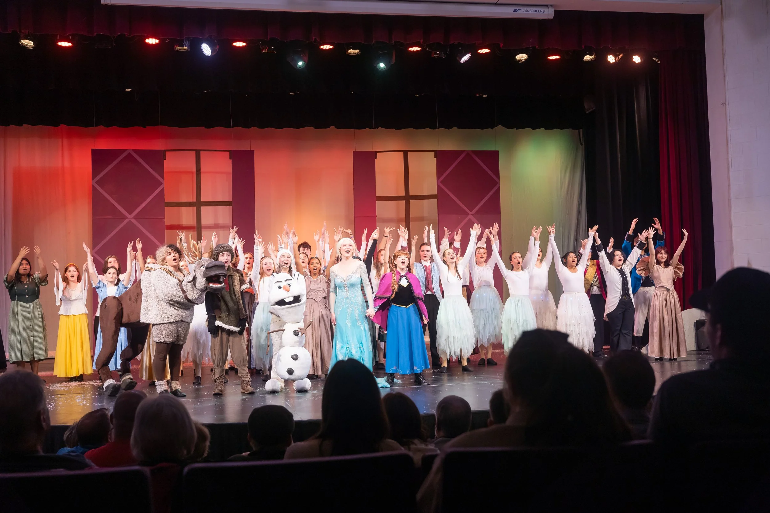 A large group of children and adults on stage, dressed in costumes from the musical 'Frozen', celebrating at the end of a performance.