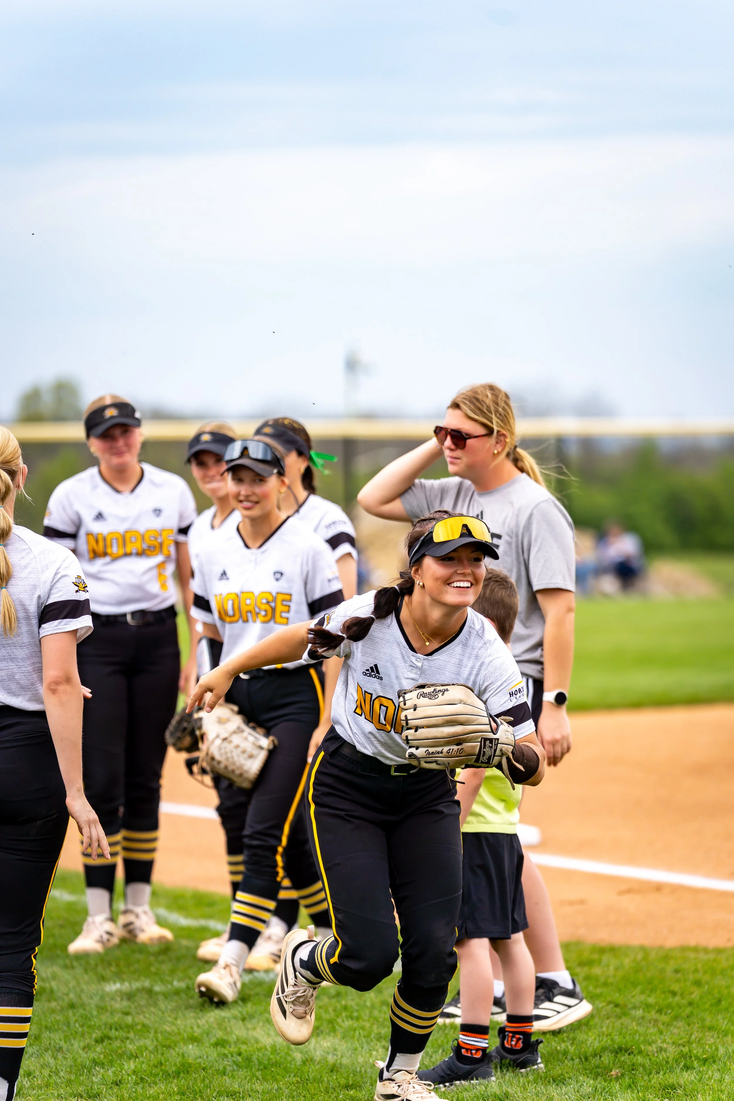 Softball players in uniform on the field, some smiling, participating in a pre-game or warm-up activity during daytime.