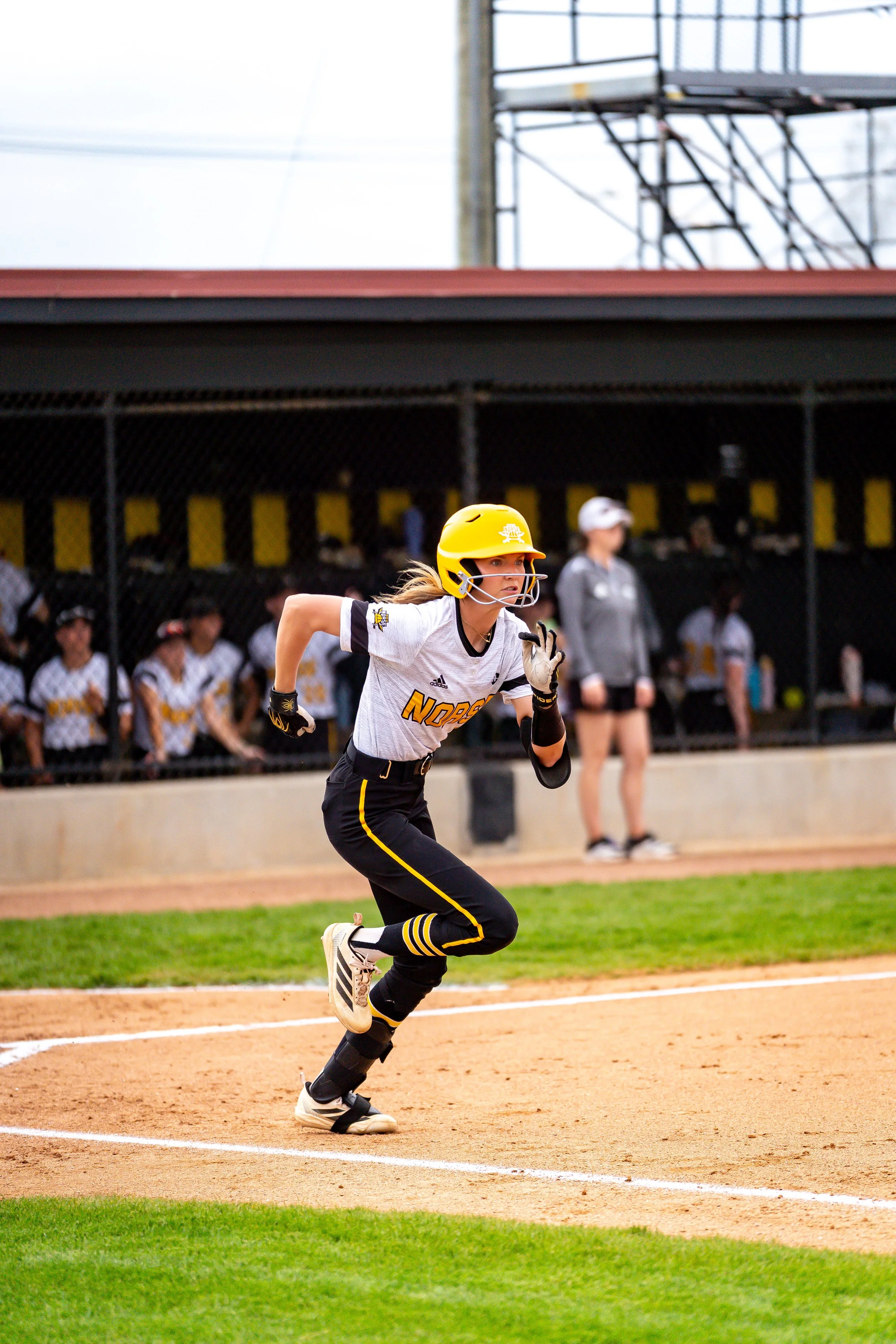 A female softball player in a yellow helmet and black pants running around bases during a game, with her team and coaches watching from the dugout in the background.