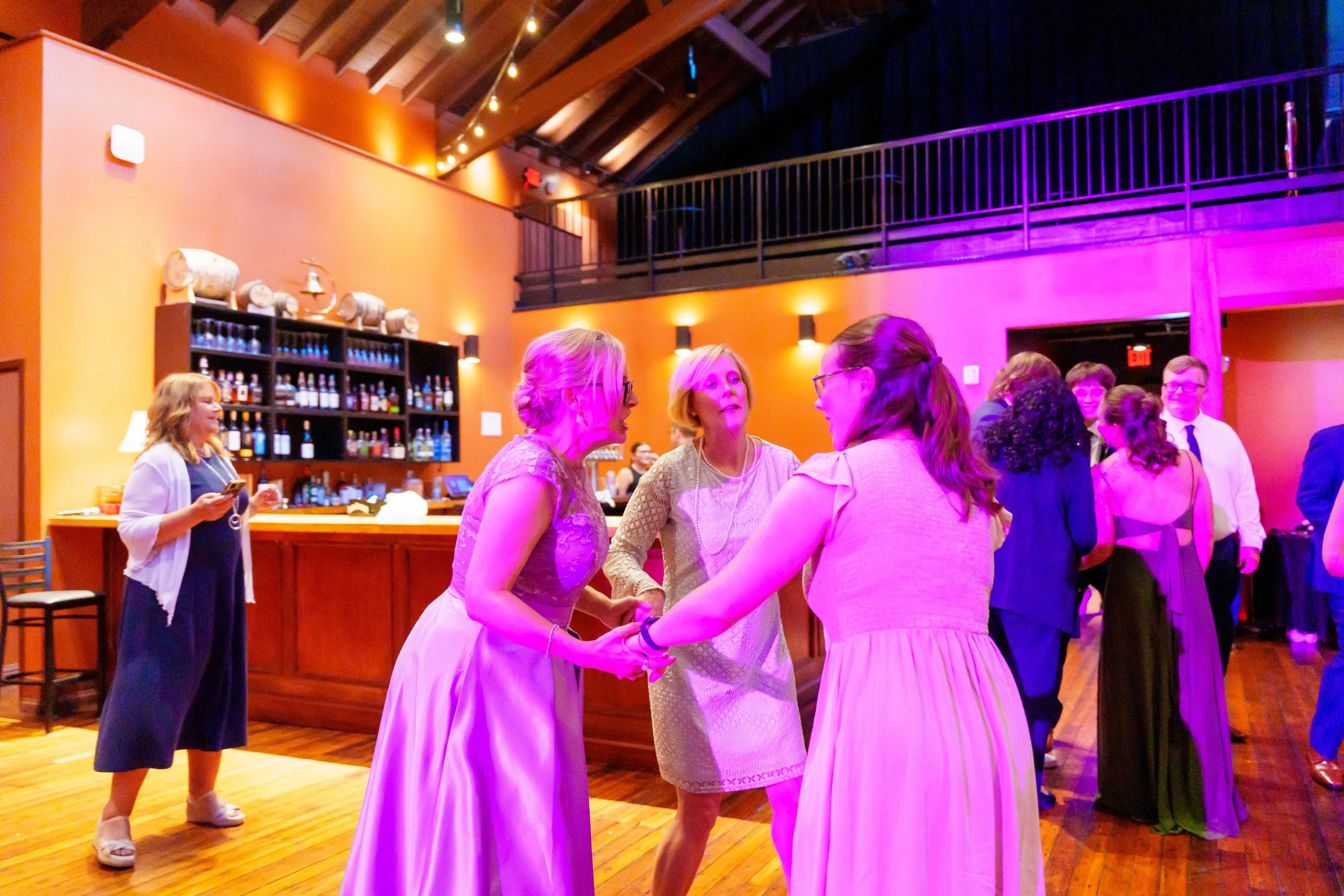 People dancing at a social event in a warmly lit venue with orange walls and wooden floors, with a bar and shelves of liquor bottles in the background.