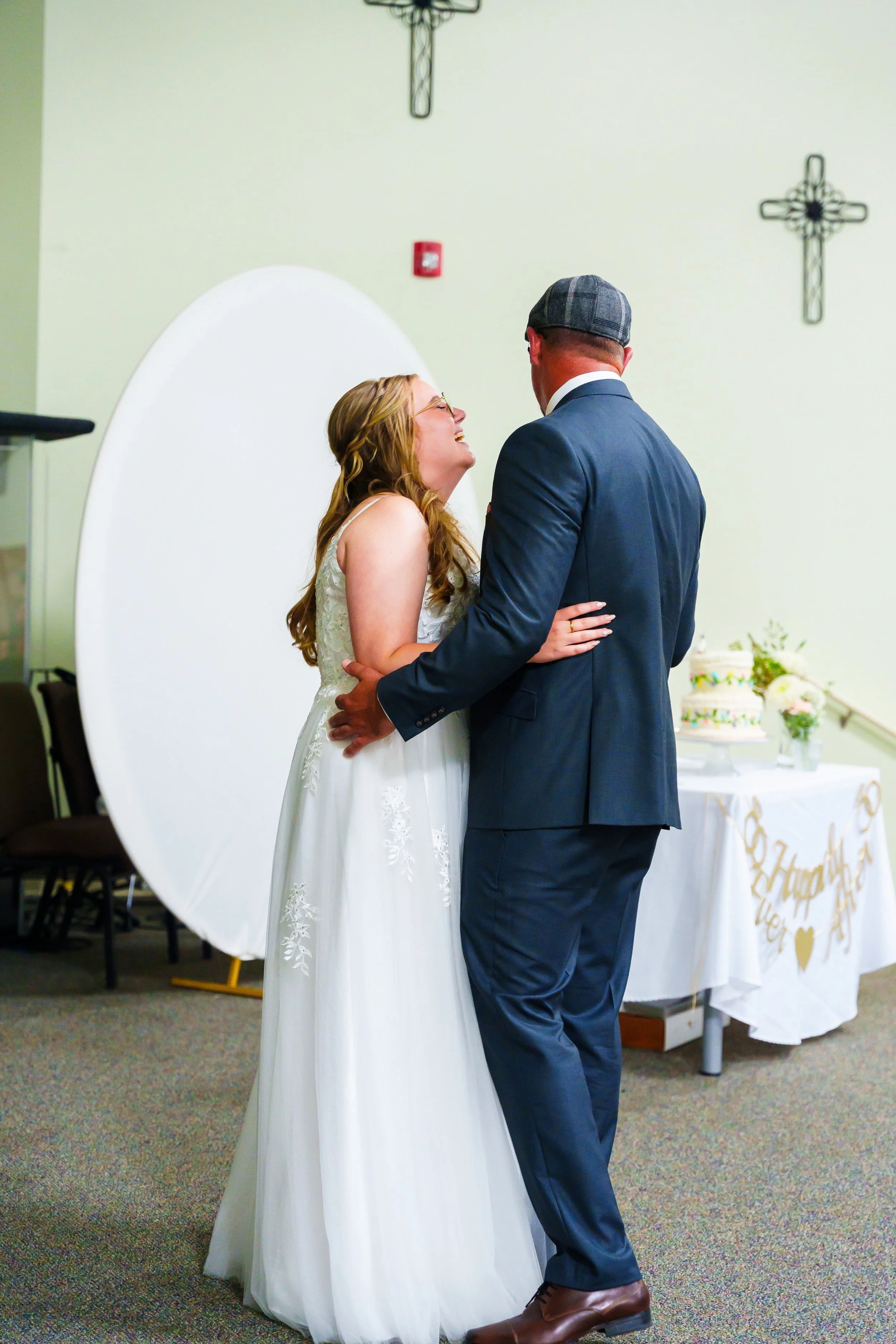 A bride and groom sharing a dance at their wedding reception, with the bride wearing a white wedding gown and the groom dressed in a navy suit and a gray hat. There are Christian crosses on the wall behind them and wedding decorations on a table near