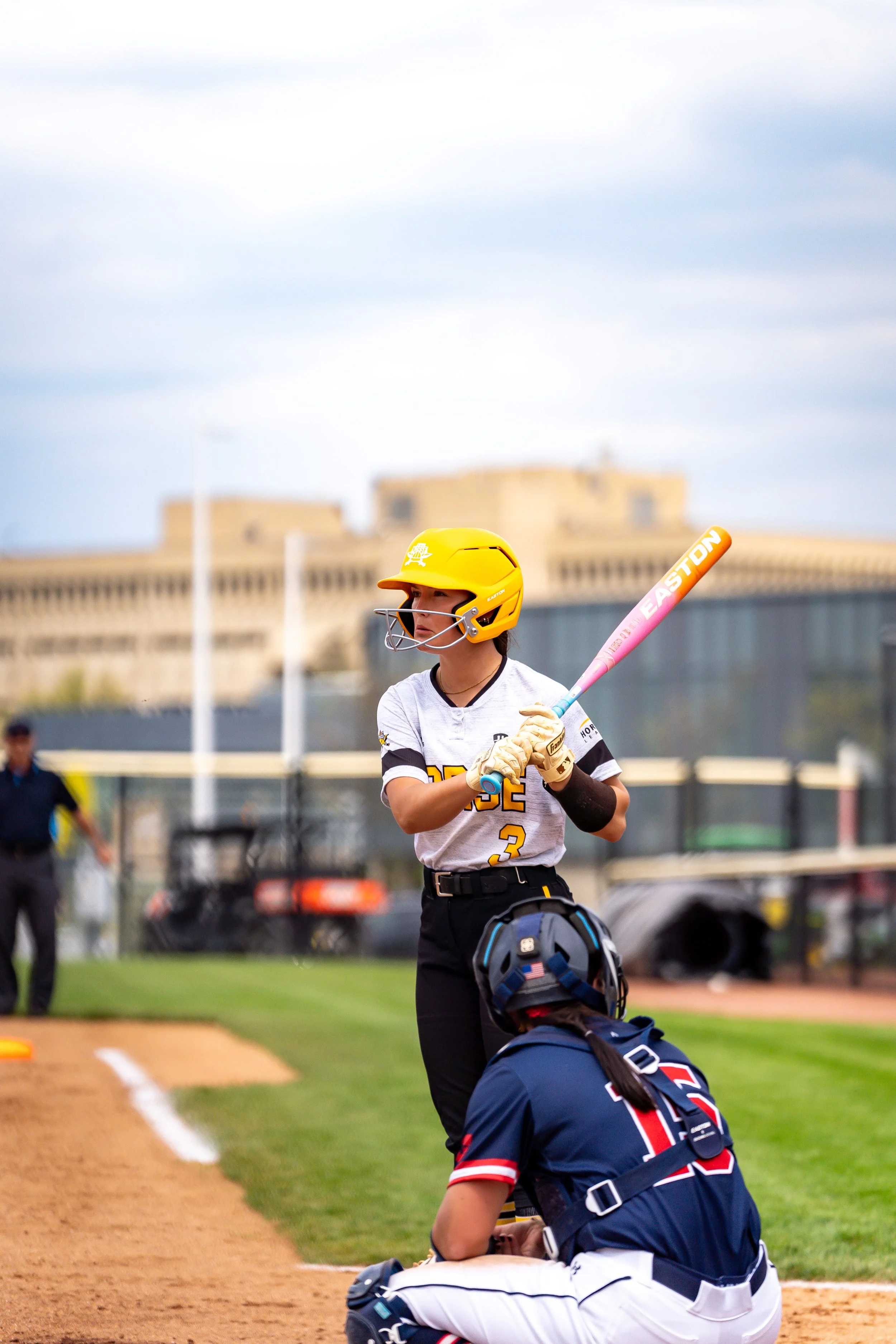 A baseball player wearing a yellow helmet, white shirt with the number 3, holding a pink Easton bat, preparing to bat during a game.