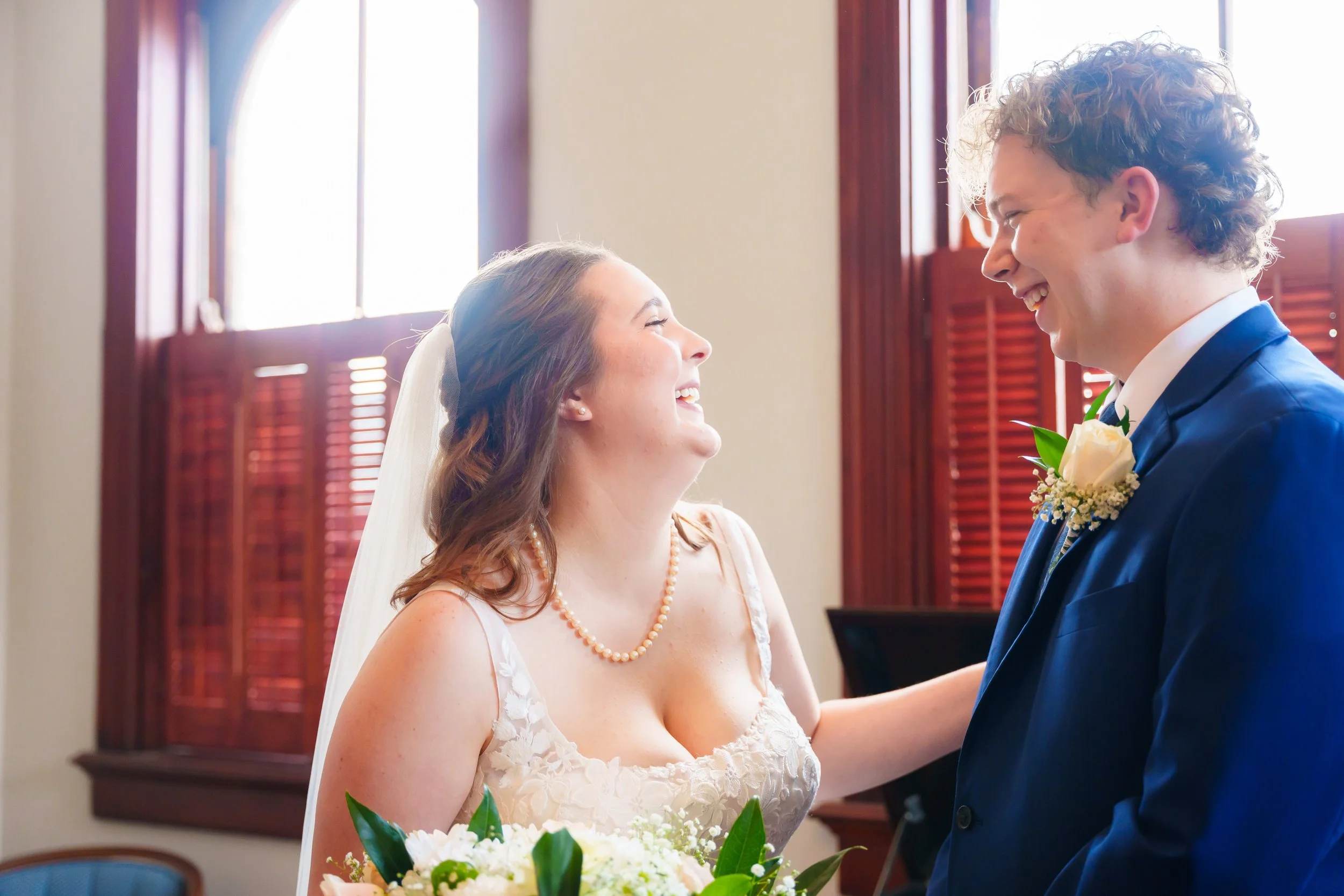 A bride and groom smiling at each other during their wedding ceremony inside a room with wooden window shutters.