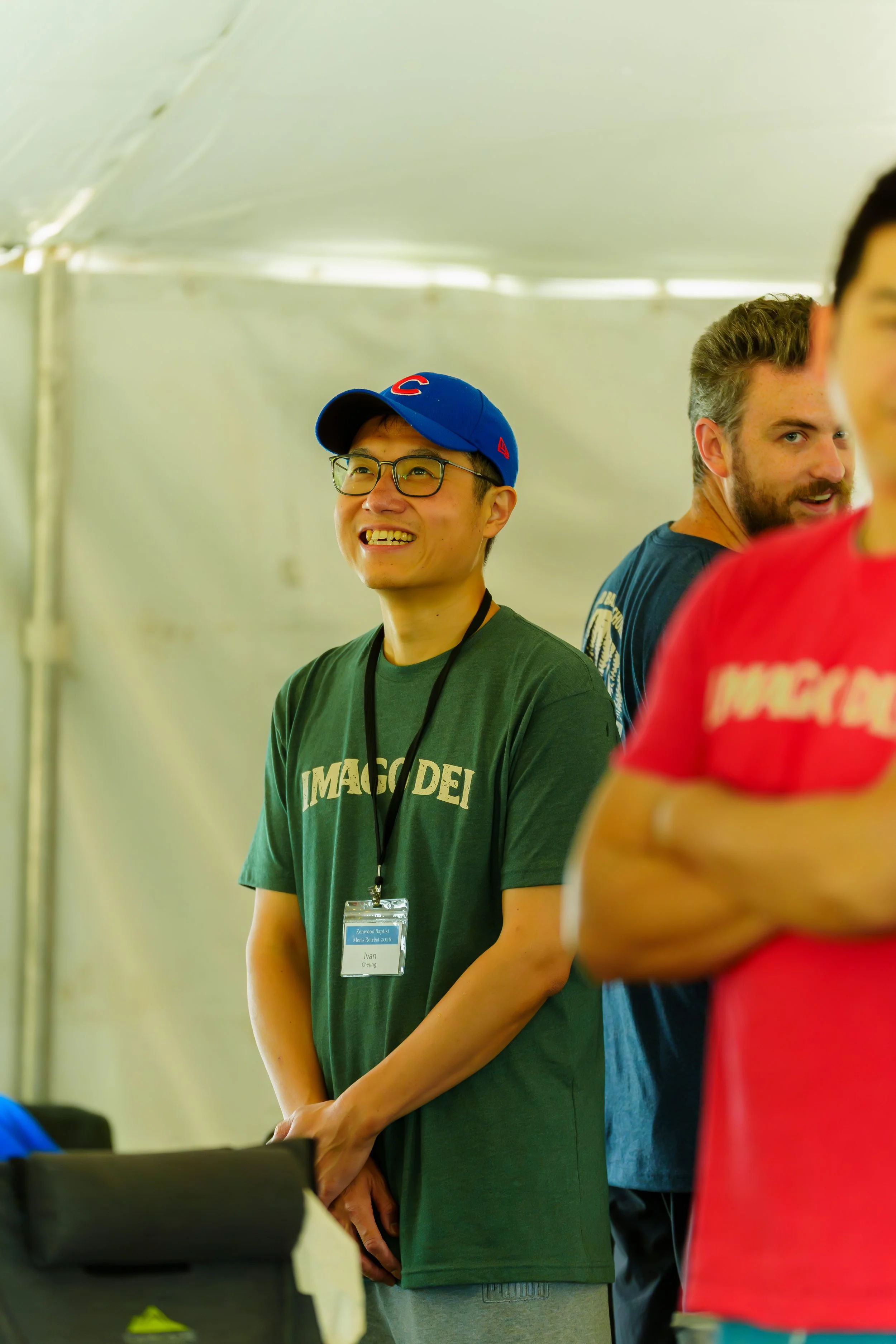 A group of people indoors, with two individuals prominently visible: a young man wearing glasses and a blue baseball cap, smiling, and another man with a beard and tousled hair, looking to the side. The young man is wearing a green shirt with white t