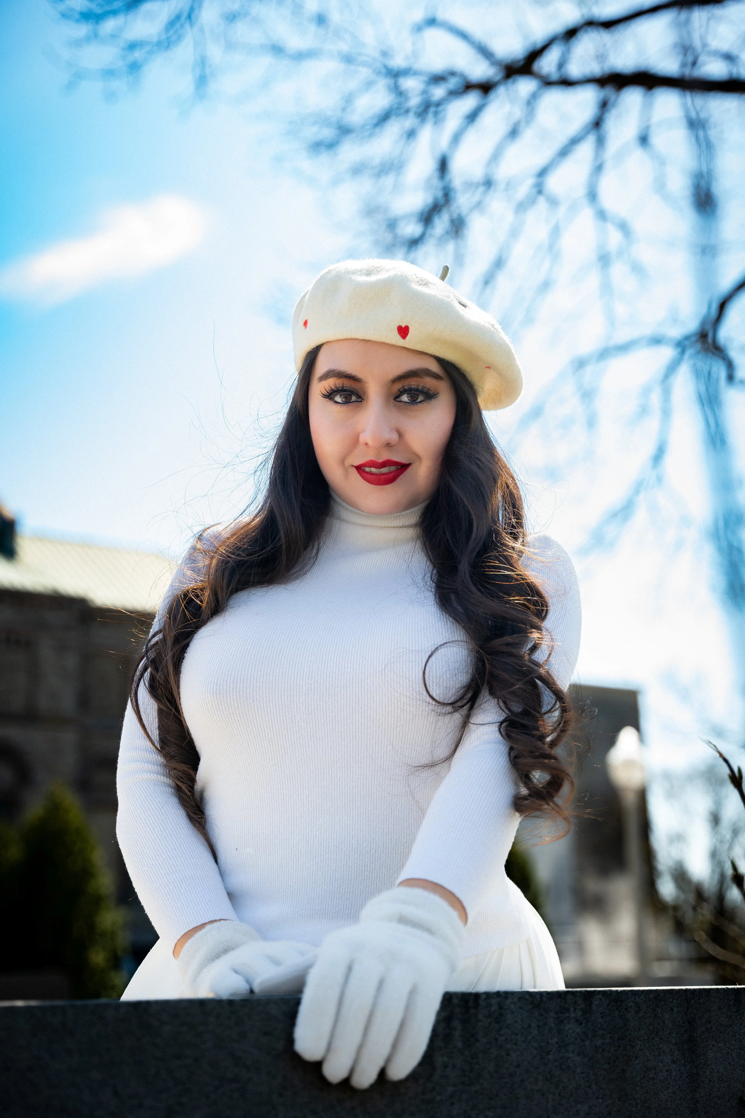 A woman with long dark hair, wearing a white beret with red hearts, a white turtleneck sweater, and white gloves, standing outdoors on a sunny day.