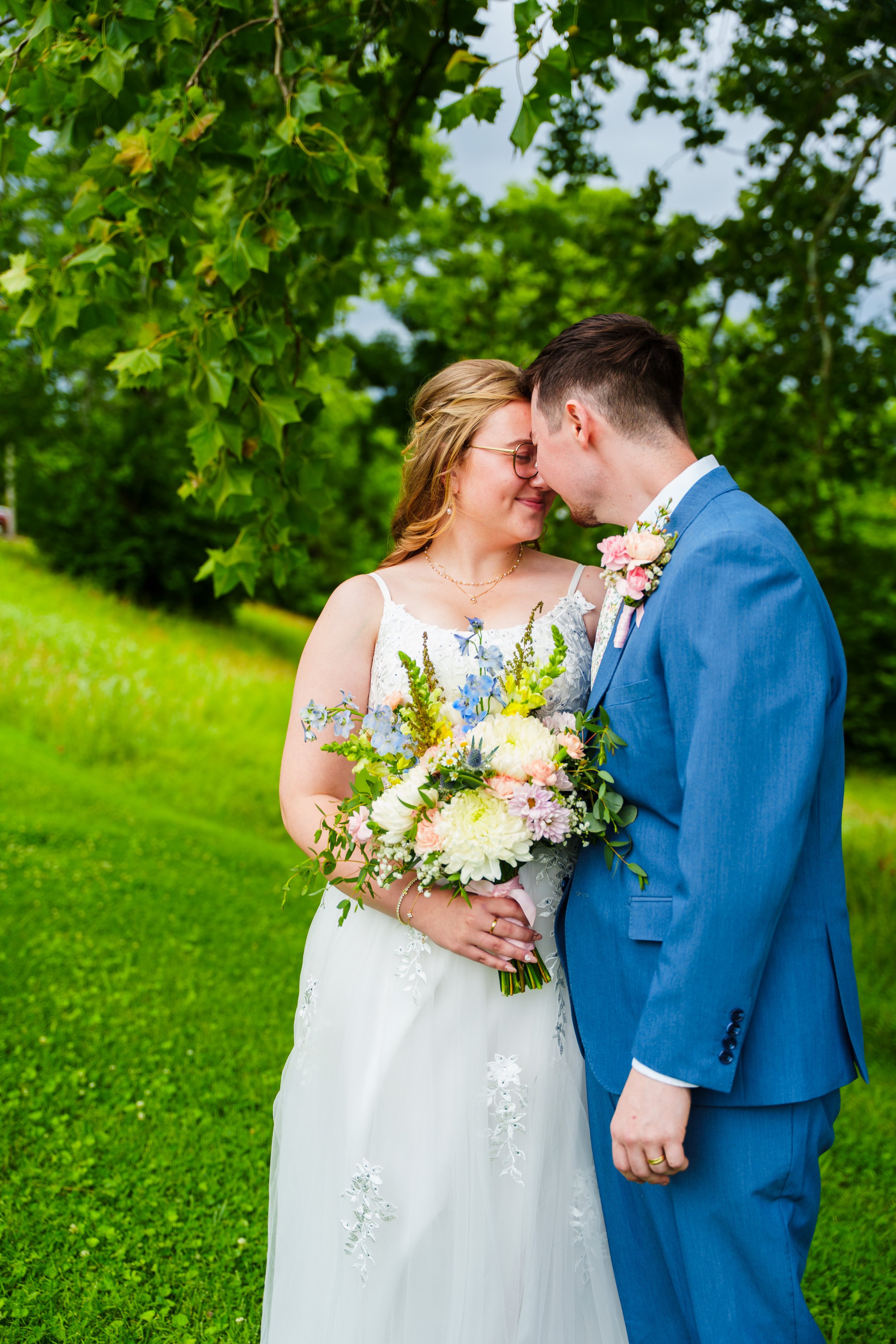 A bride and groom holding hands and touching foreheads outdoors, surrounded by green trees and grass, on a cloudy day.