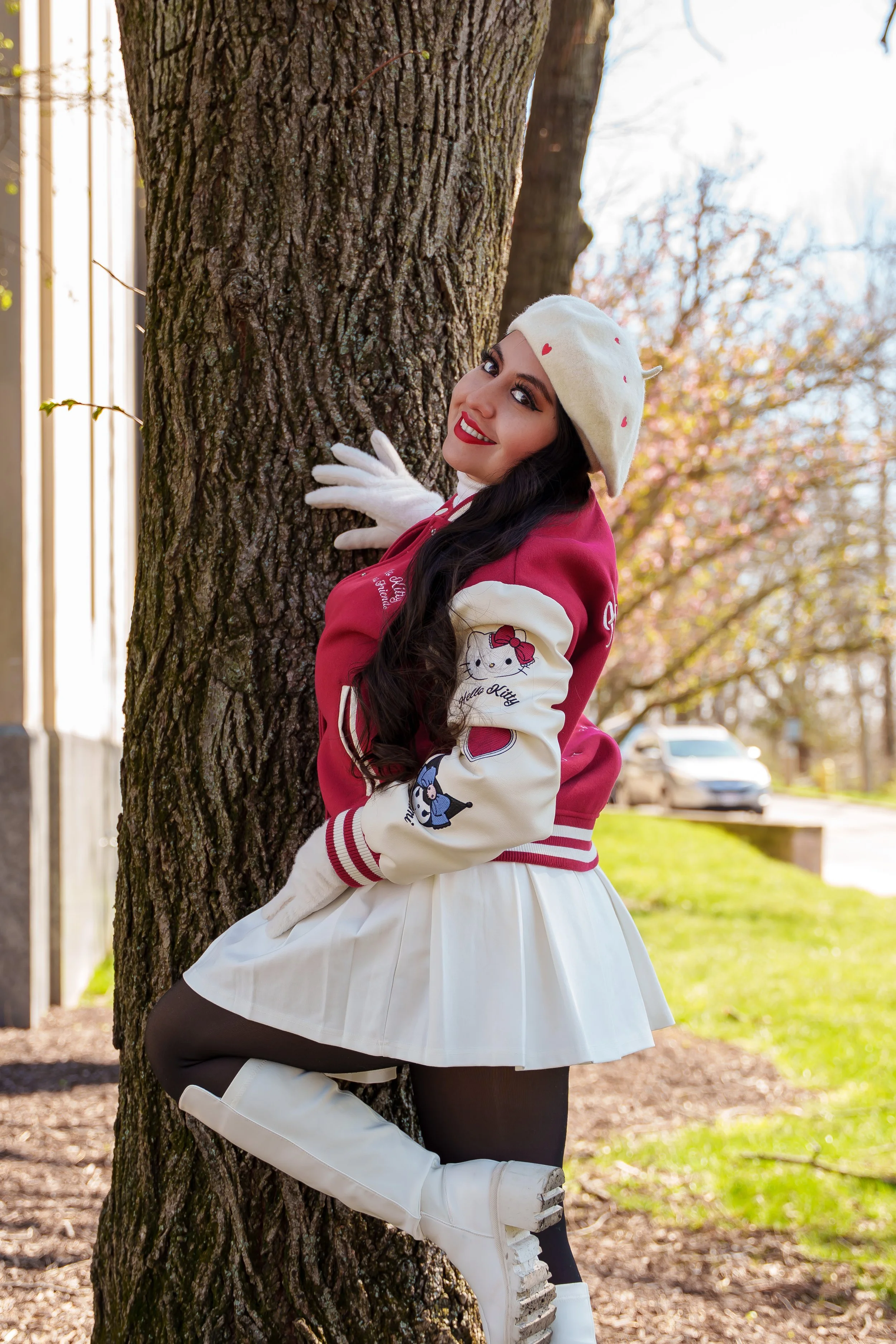 A woman with long dark hair and bright red lipstick posing outdoors next to a tree. She is wearing a white beret with red dots, a red and white letterman jacket with Hello Kitty patches, a white skirt, black tights, and white knee-high boots, smiling