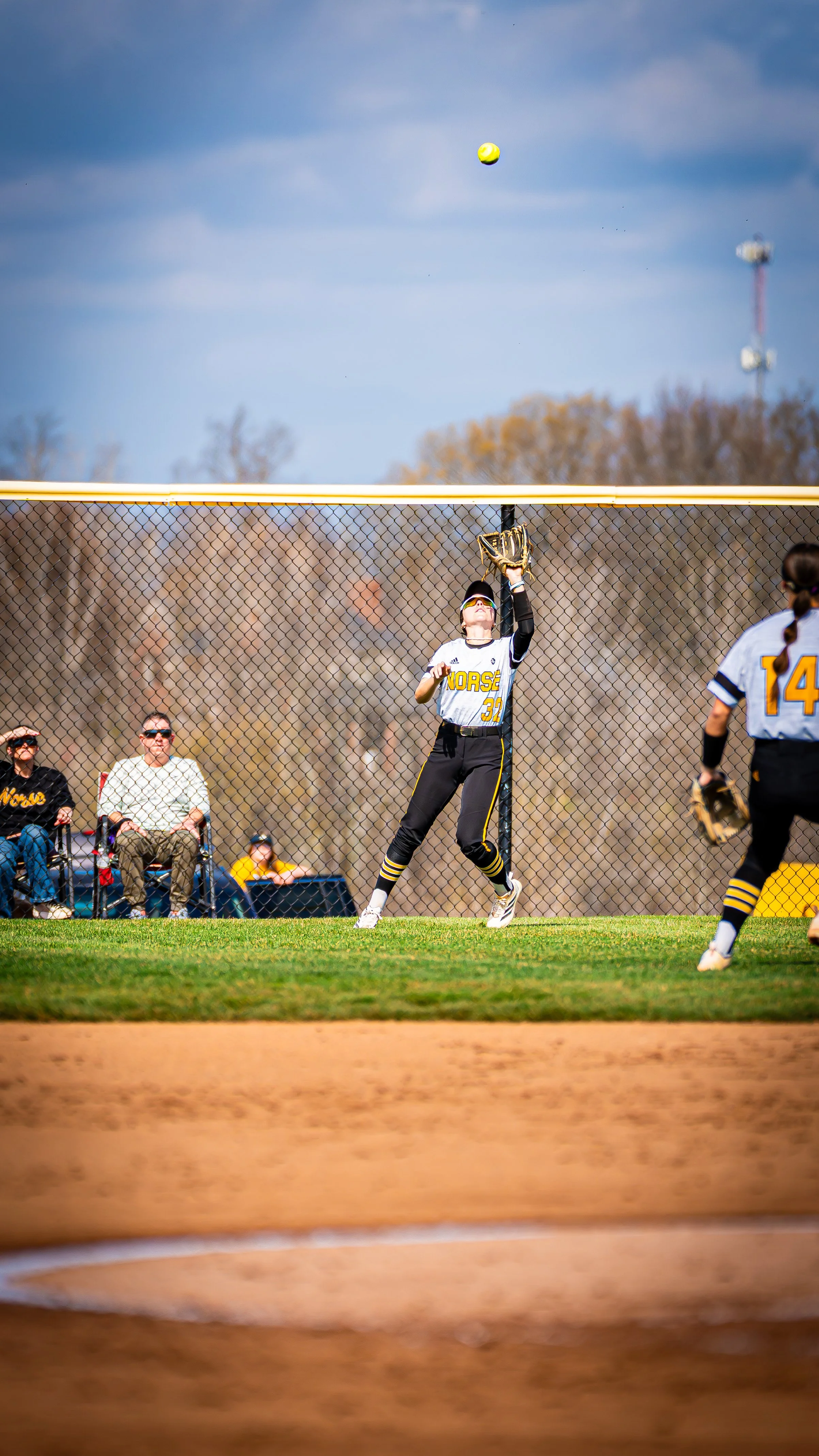 A softball player in a white and yellow uniform jumps to catch a ball at the outfield fence during a game on a sunny day. Other players and spectators are visible in the background.