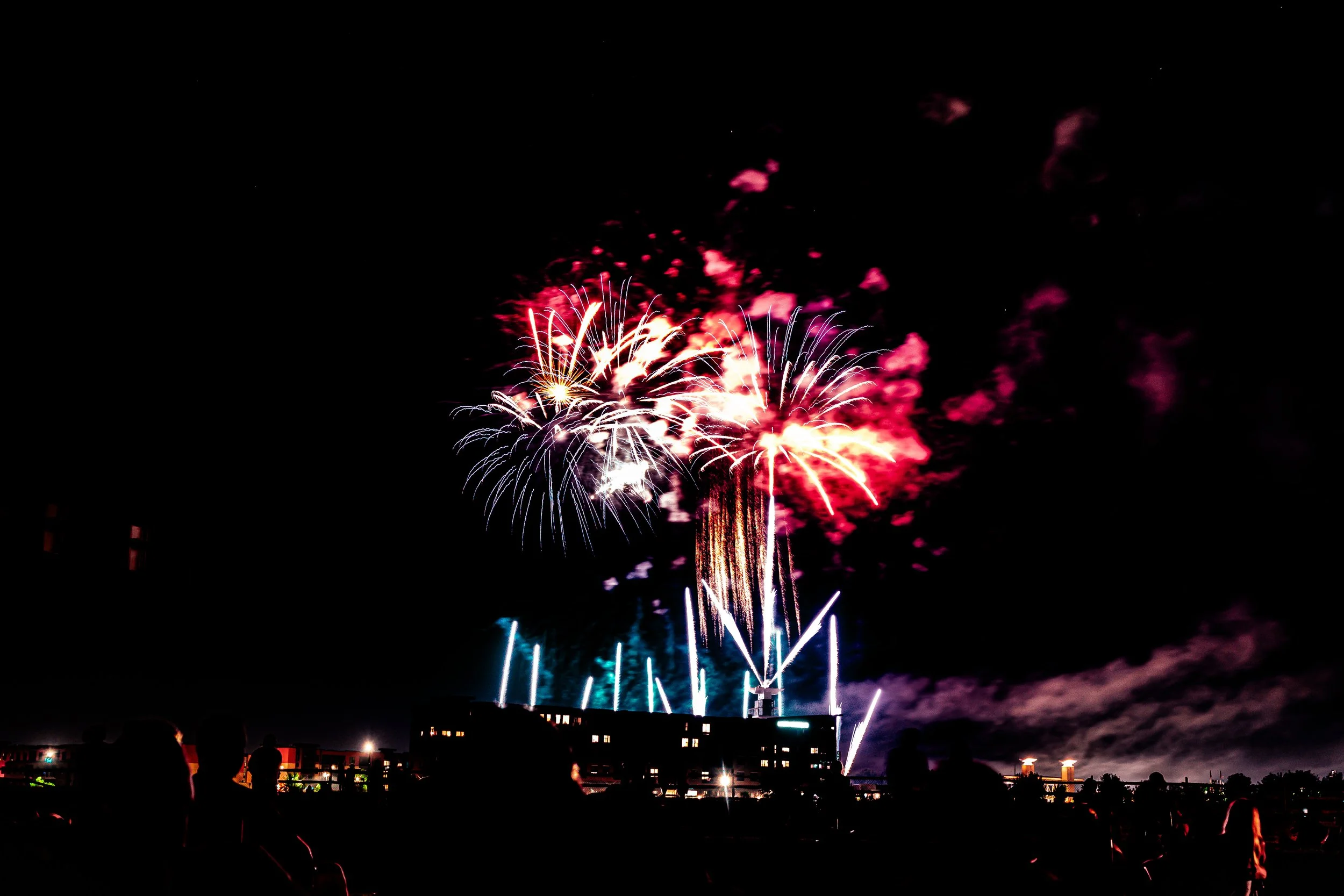Fireworks display lighting up the night sky with red, pink, and white bursts. Silhouettes of people watching in the foreground.