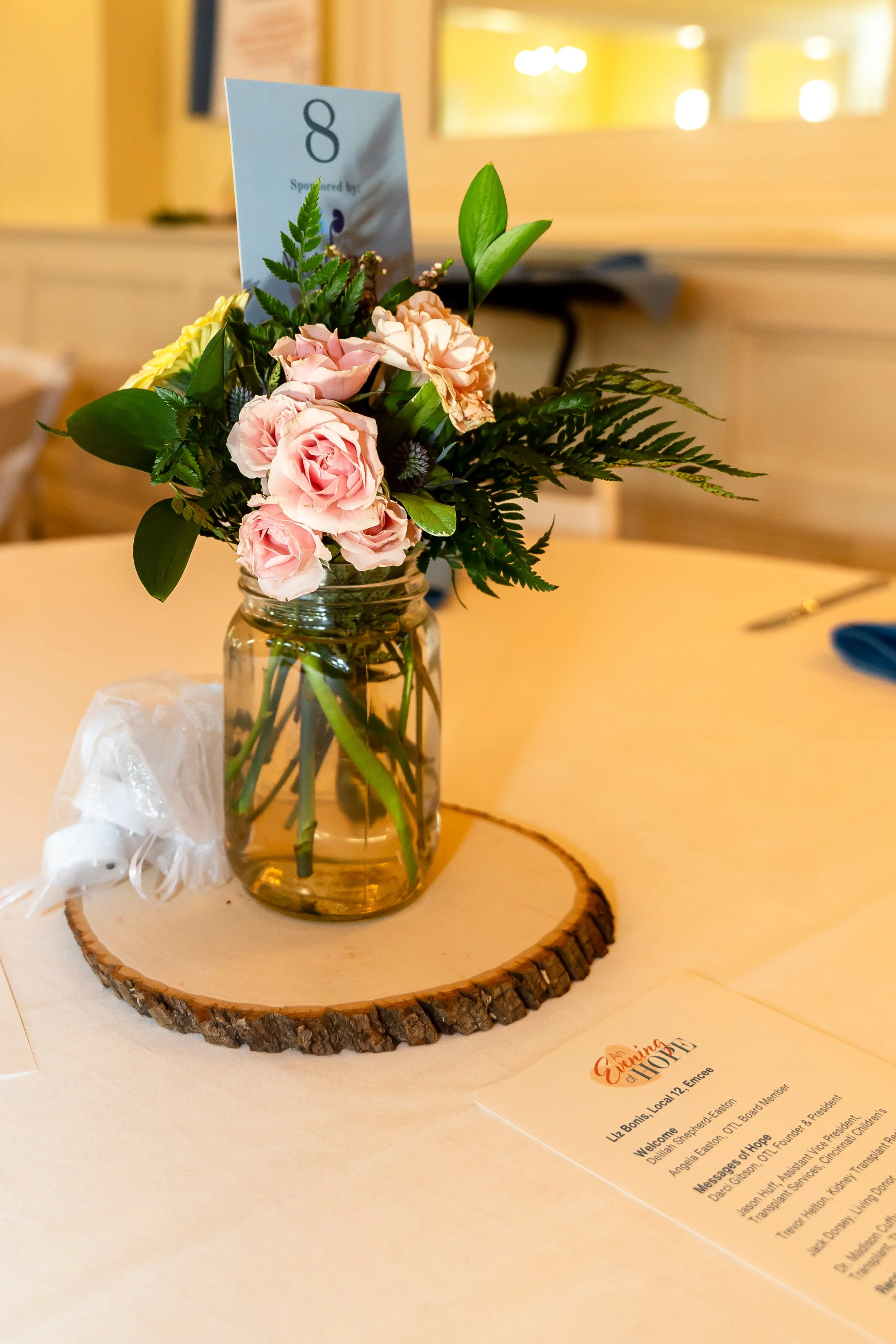 A small bouquet of pink roses, yellow flowers, and various greenery arranged in a glass jar on a wooden tree slice with a website flyer nearby.