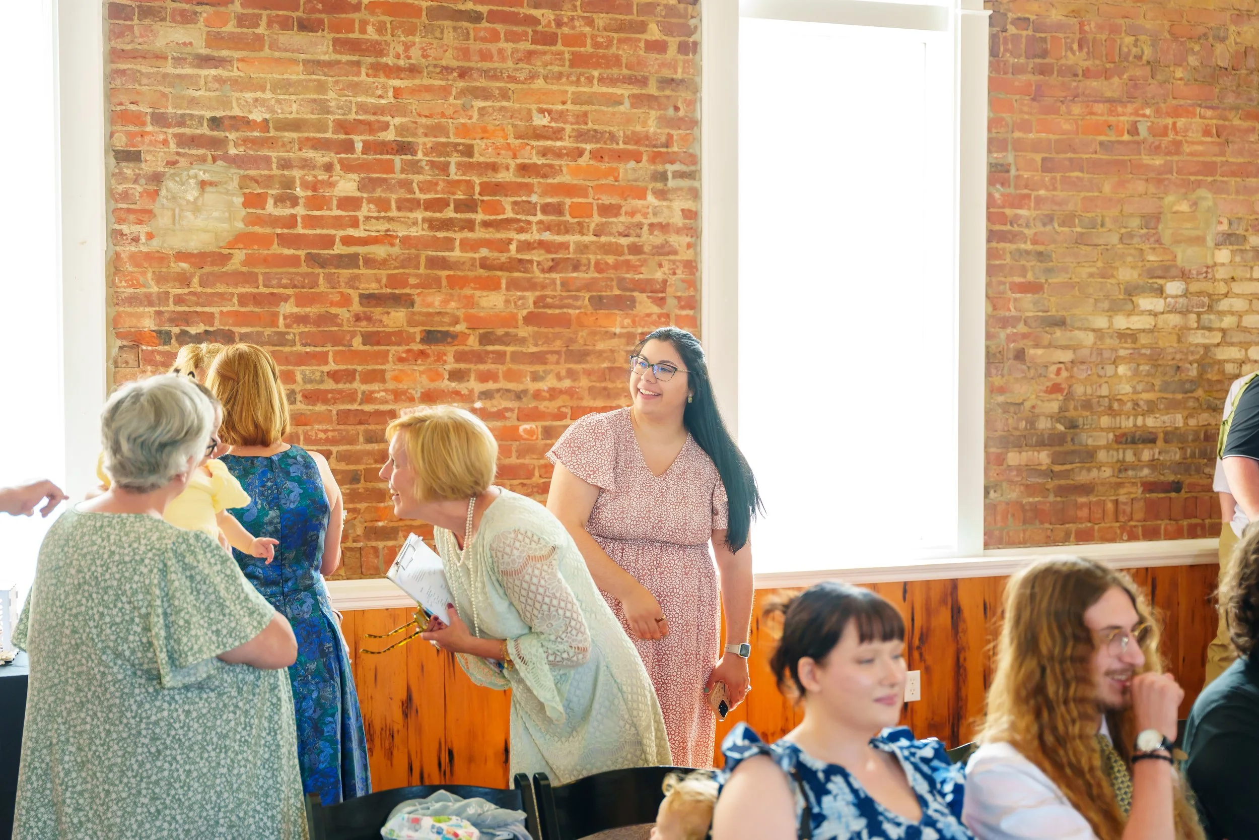 A group of women and elderly people talking and interacting in a brightly lit room with large windows, exposed brick wall, and wooden paneling. Some women are smiling and holding papers, engaged in conversation.