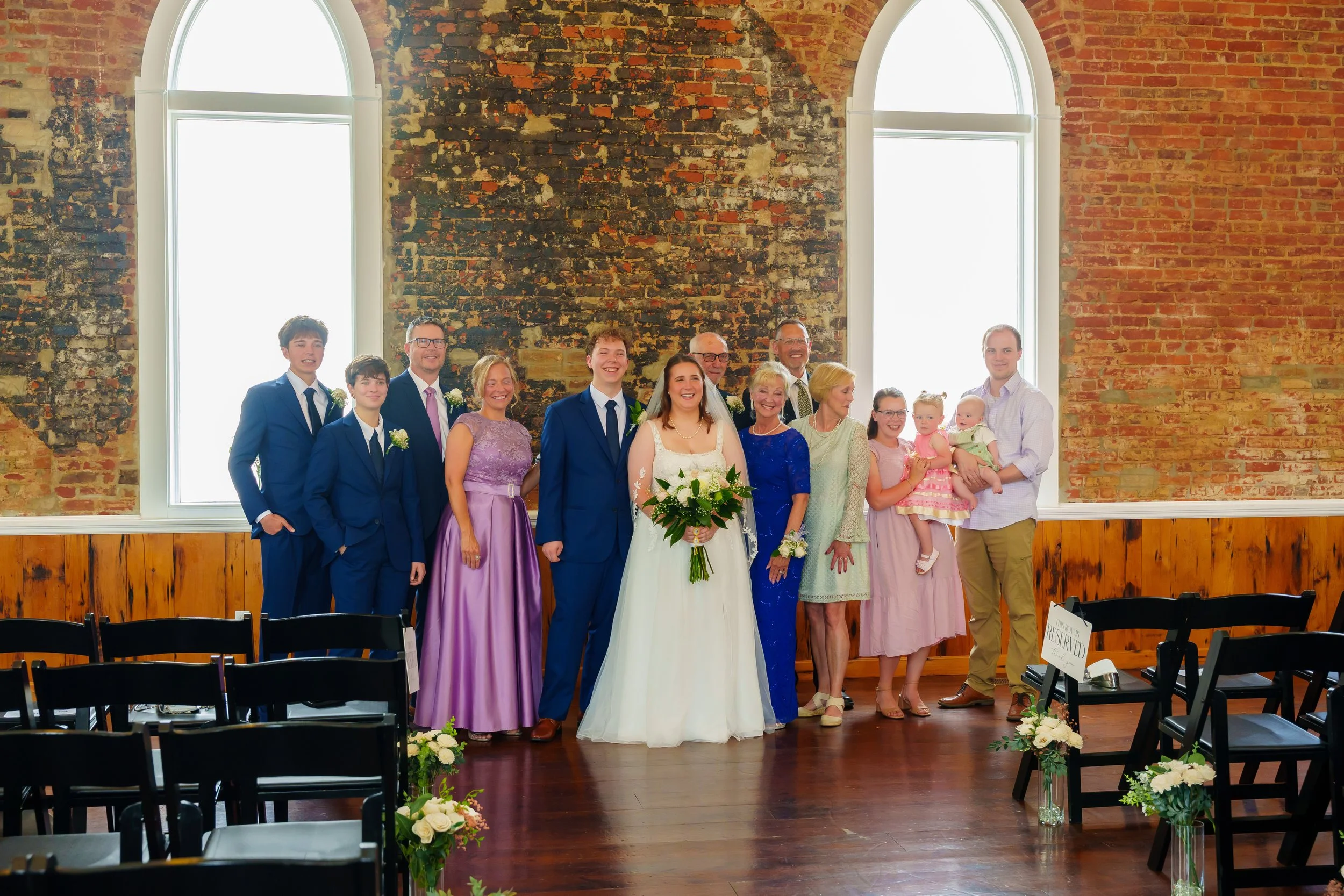 Group of people at a wedding celebration standing together in front of large arched windows in a rustic brick-walled room. The group includes the bride wearing a white dress and veil holding a bouquet, and several men and women dressed in formal atti