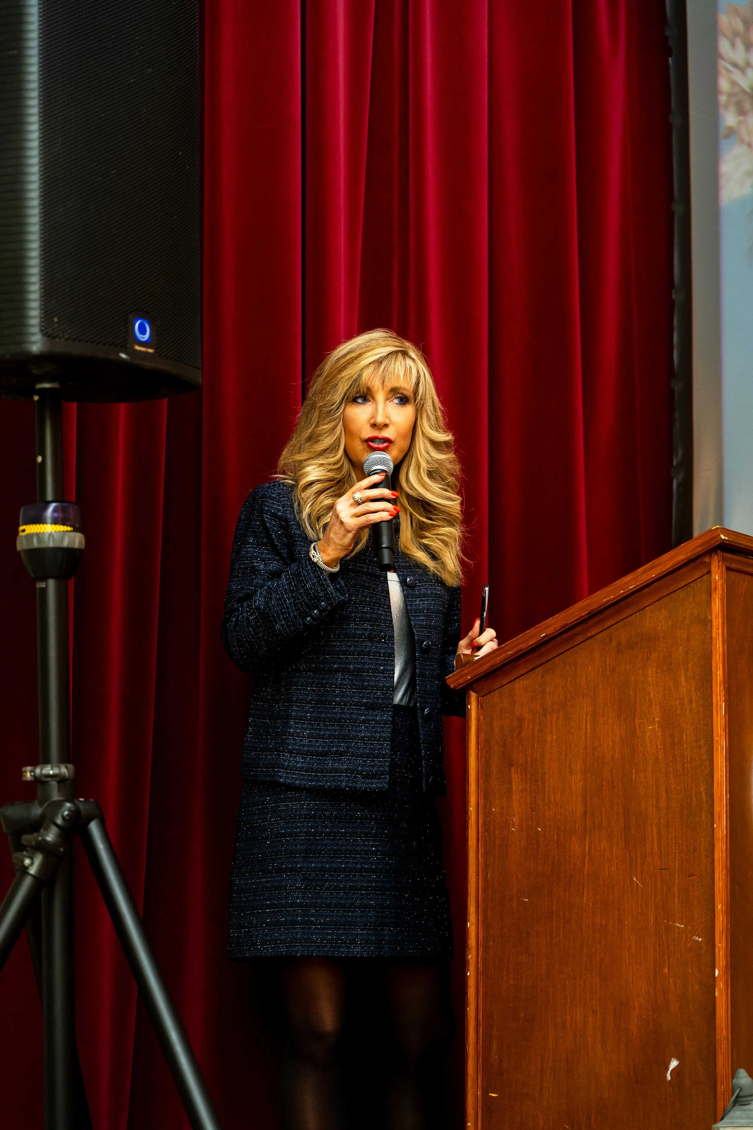 A woman with long blonde hair, wearing a dark dress, speaking into a microphone from a podium in front of red curtains.