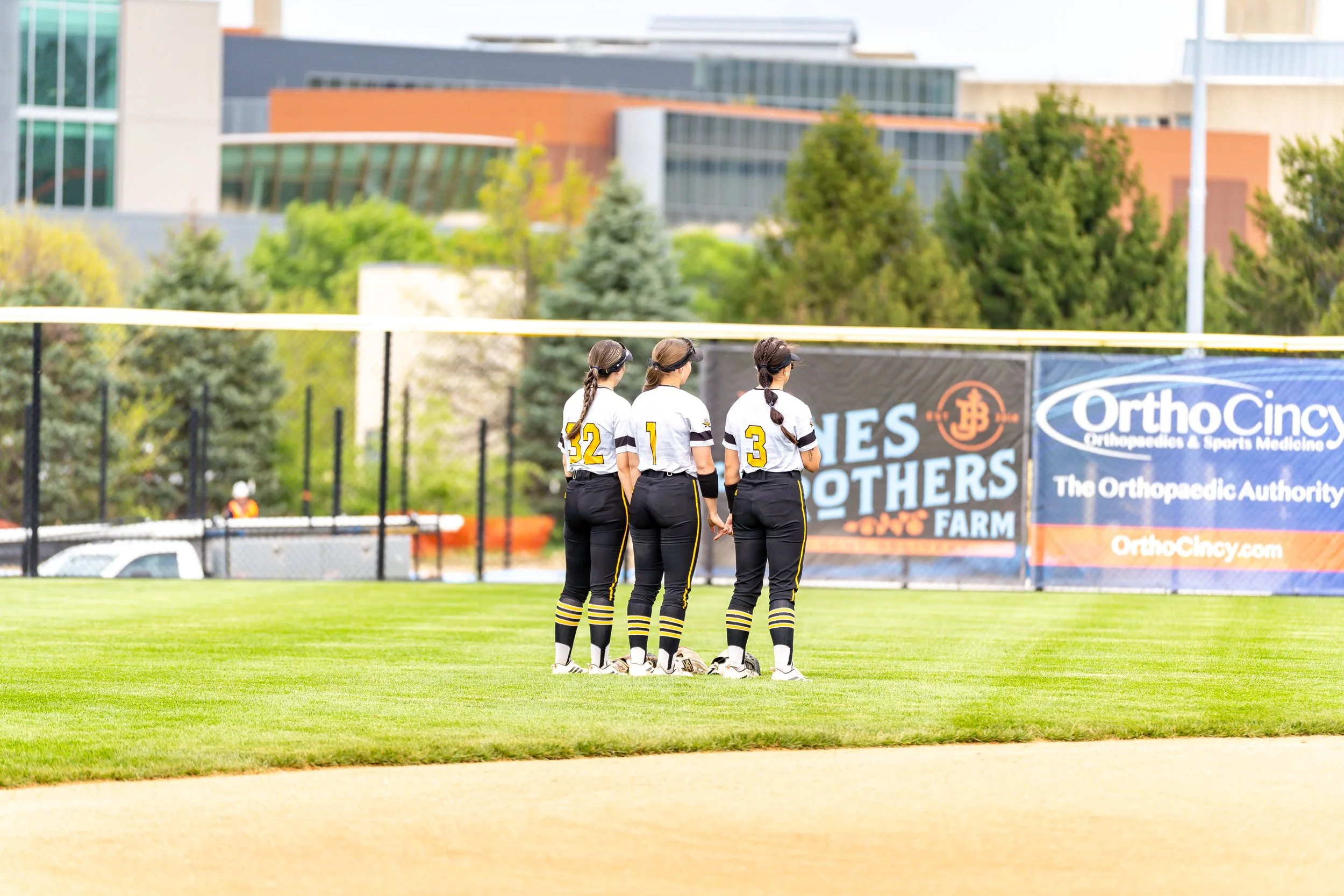Four female softball players stand by the field, facing away, wearing black and yellow uniforms with numbers 32, 1, and 3. They are watching a game, with banners and trees in the background.