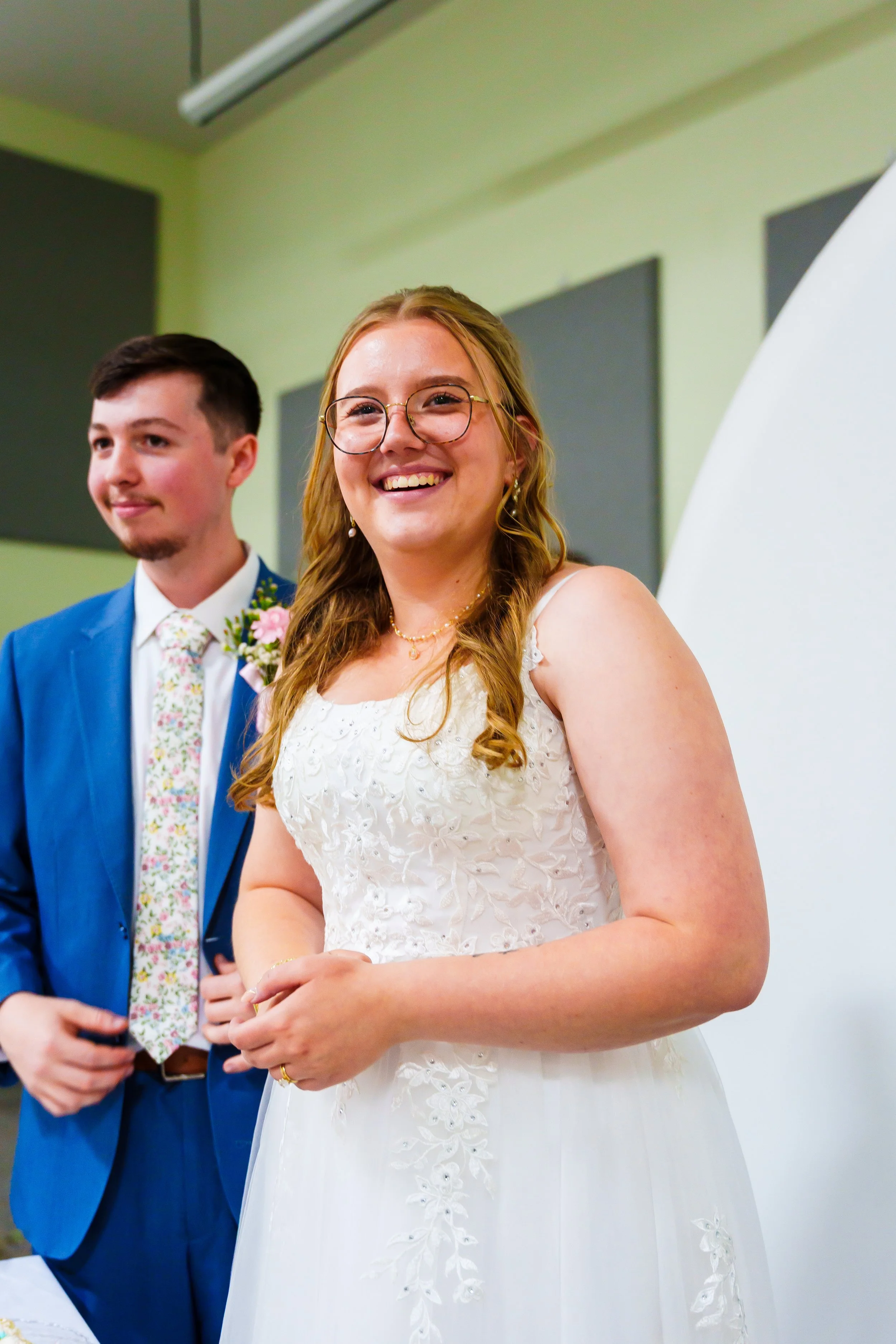 A woman in a white wedding dress and glasses smiling, standing next to a man in a blue suit with a floral tie, during a wedding ceremony.