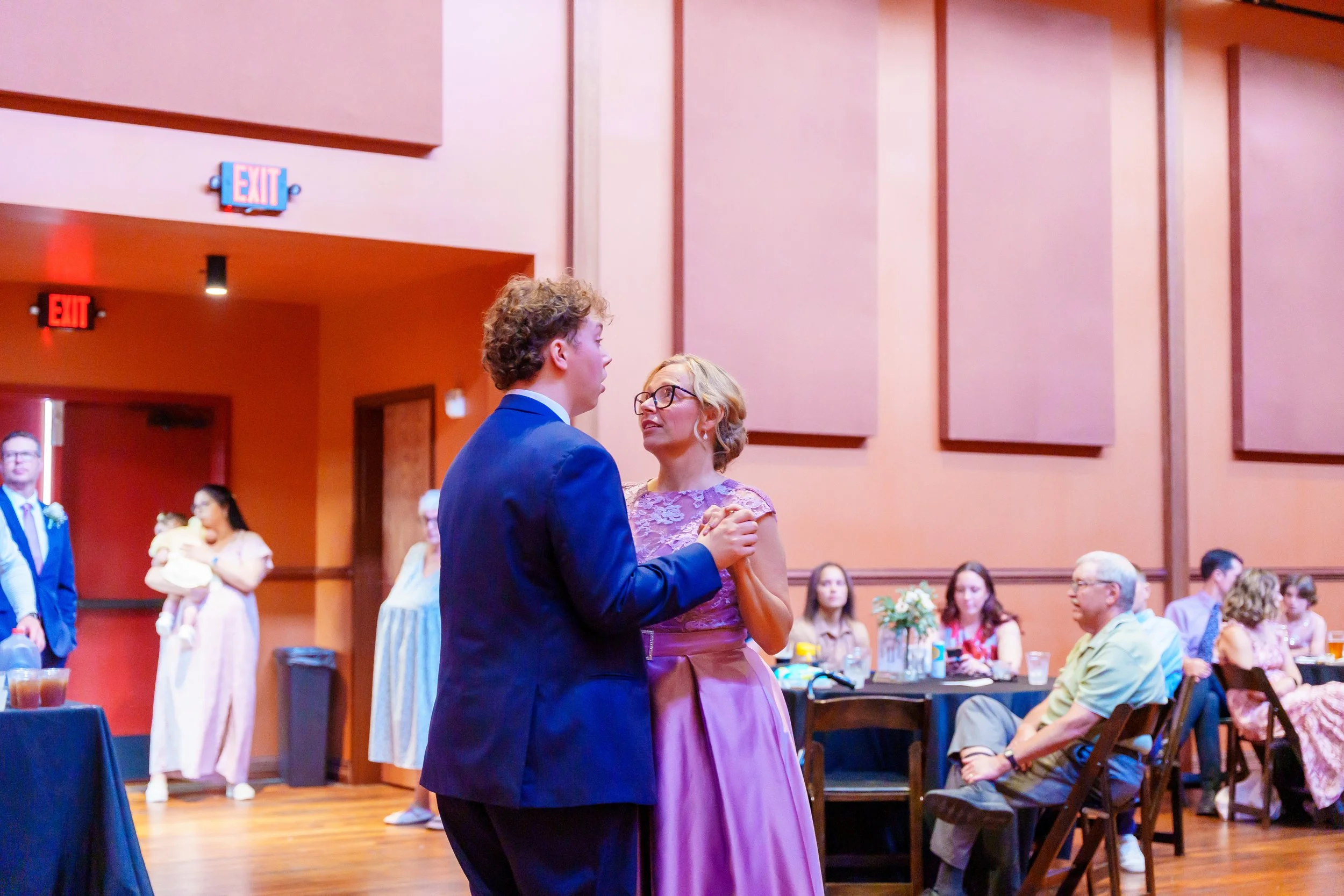 A young man in a blue suit and a middle-aged woman in a purple dress dance together at a formal event in a large room with pink walls and wood paneling. There are people seated at tables and a few standing in the background, with a woman holding a ba