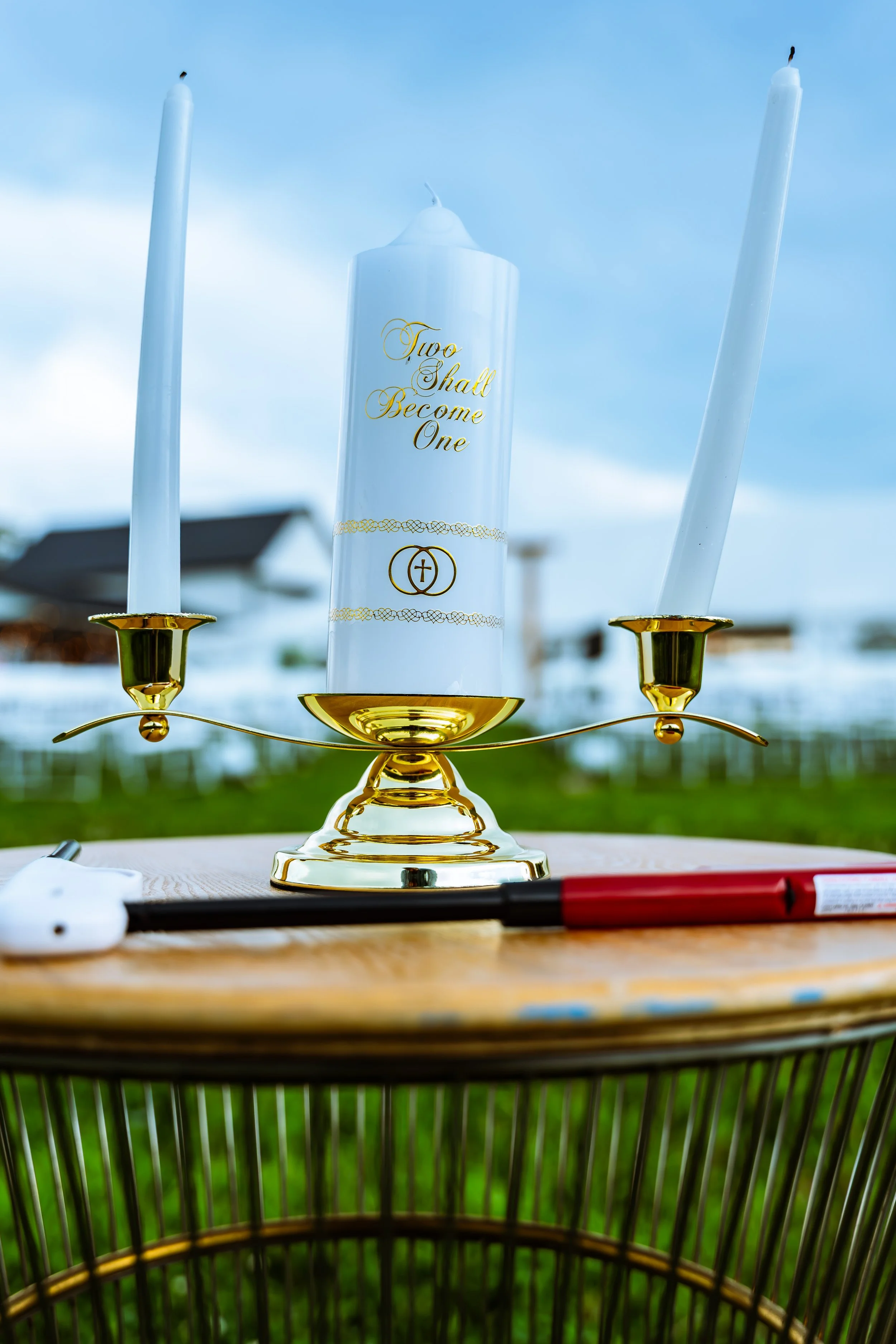 A gold candle holder with two white candles and a large white candle in the center with gold text that reads "Two Shall Become One." The background shows a blue sky with some clouds, and the candle holder is placed on a wooden surface.
