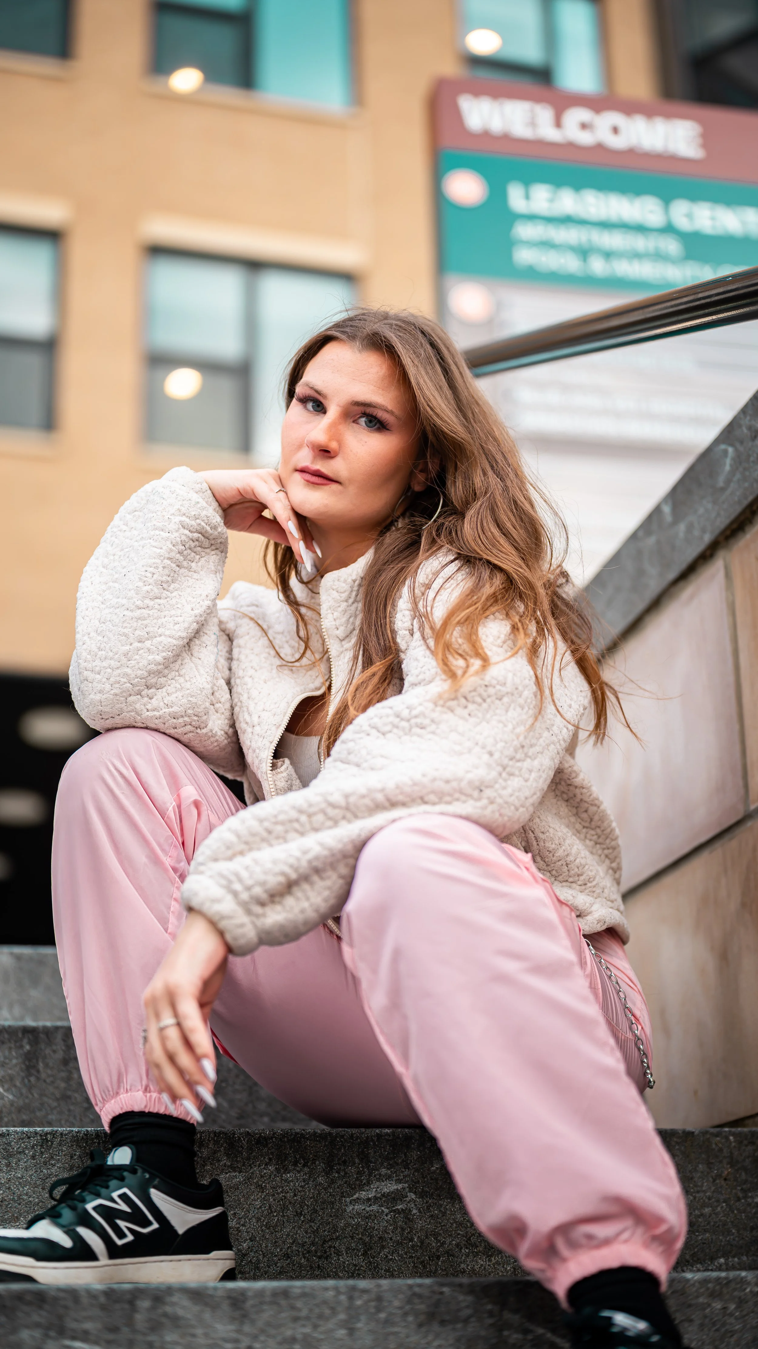 A young woman with long, wavy hair, wearing a beige fleece jacket, pink pants, and black sneakers, sitting on outdoor staircase with her legs apart and resting her chin on her hand, looking at the camera.