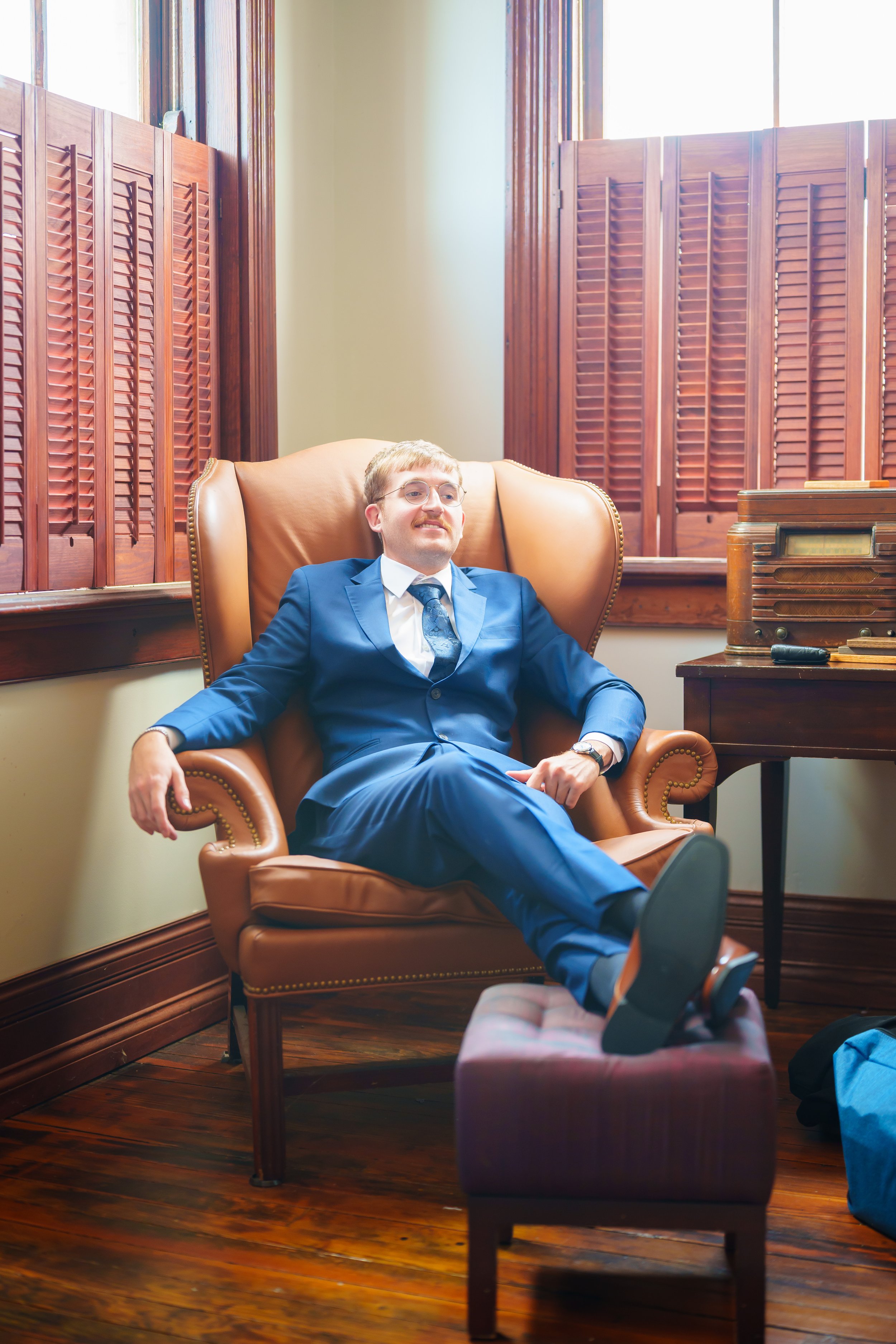A man in a blue suit and glasses with blonde hair and a mustache sitting relaxingly in a leather armchair in a room with wooden flooring, wooden window shutters, and a wooden cabinet with a vintage radio on top.