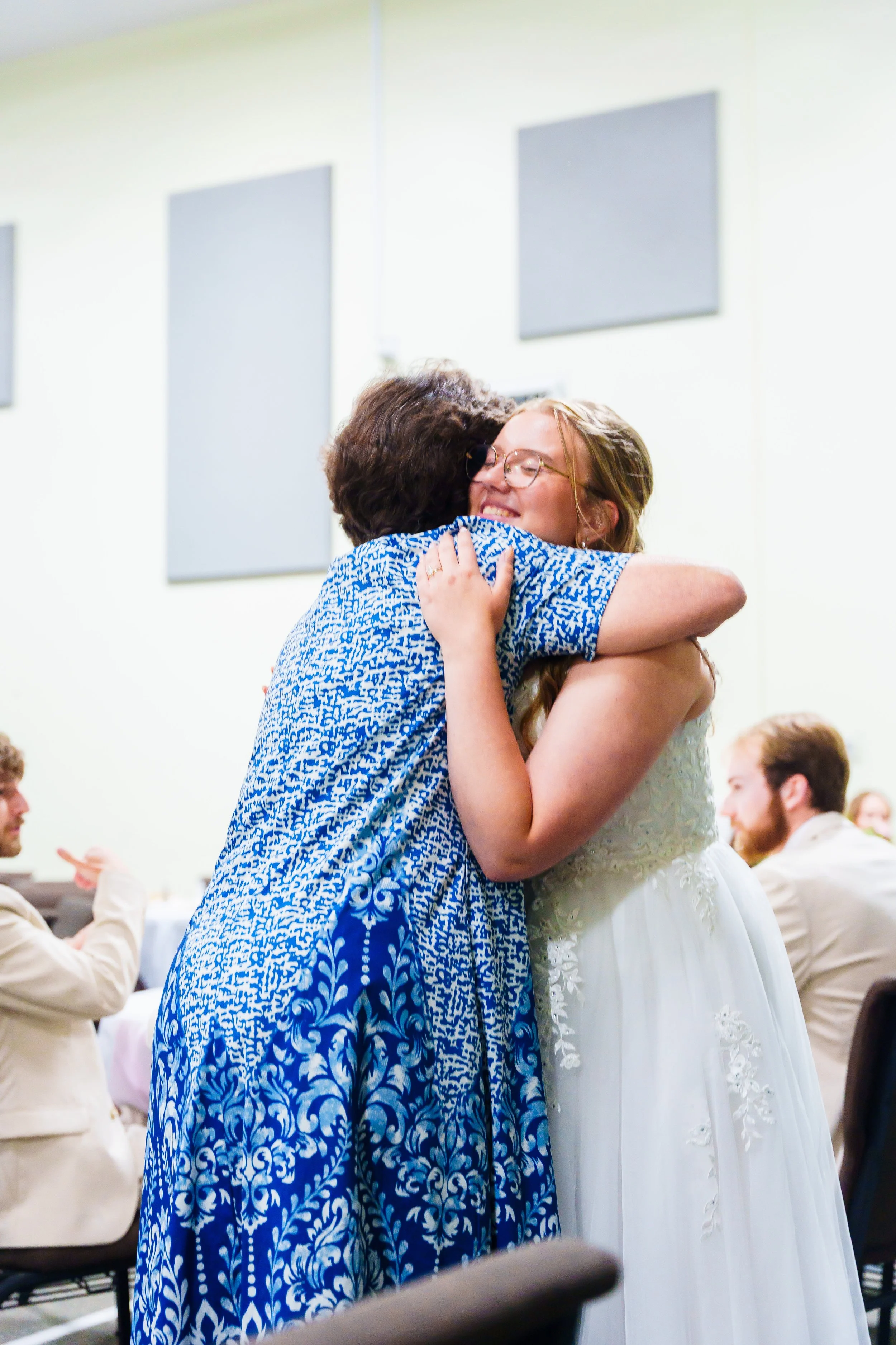 A woman in a white dress hugging a woman in a blue patterned dress, both with smiles, in a room with other people.