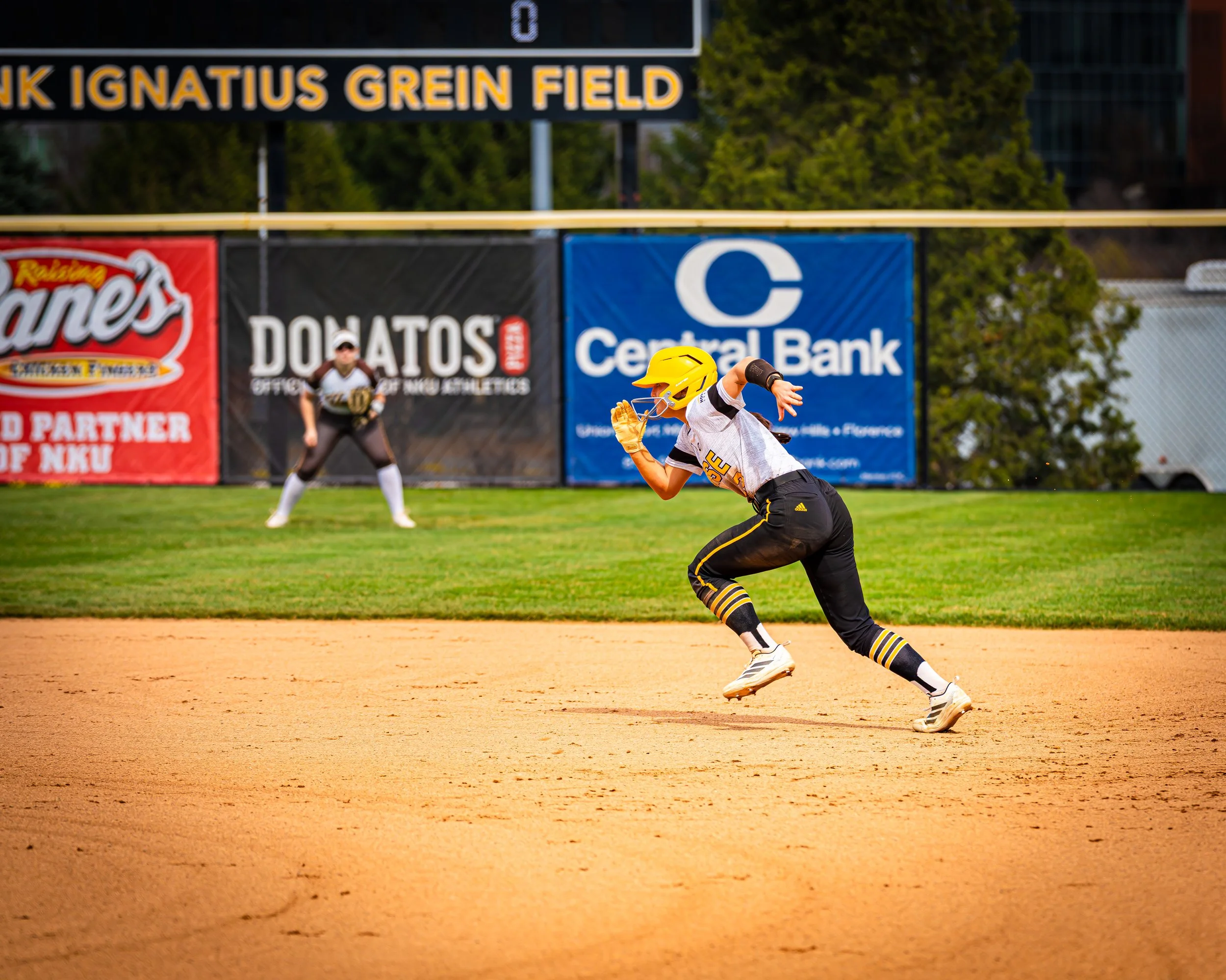 A female softball player in a white and black uniform with a yellow helmet is in the middle of a swing, against a background of a baseball field with advertising banners and a teammate or coach in the distance.