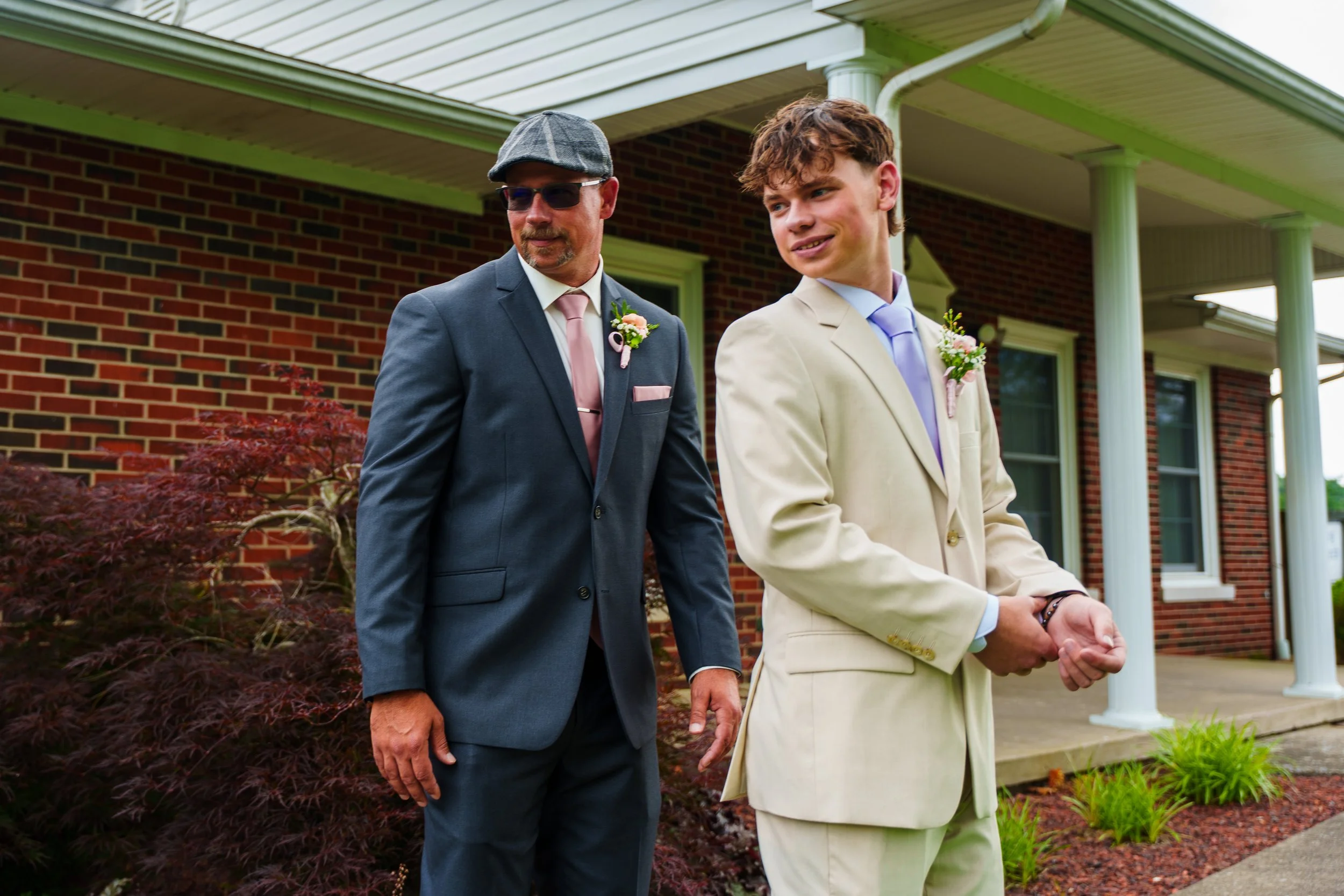 Two men in suits walking outside in front of a brick house with green trim, one older man wearing sunglasses and a flat cap, and a younger man with curly hair, both wearing boutonnières and smiling.