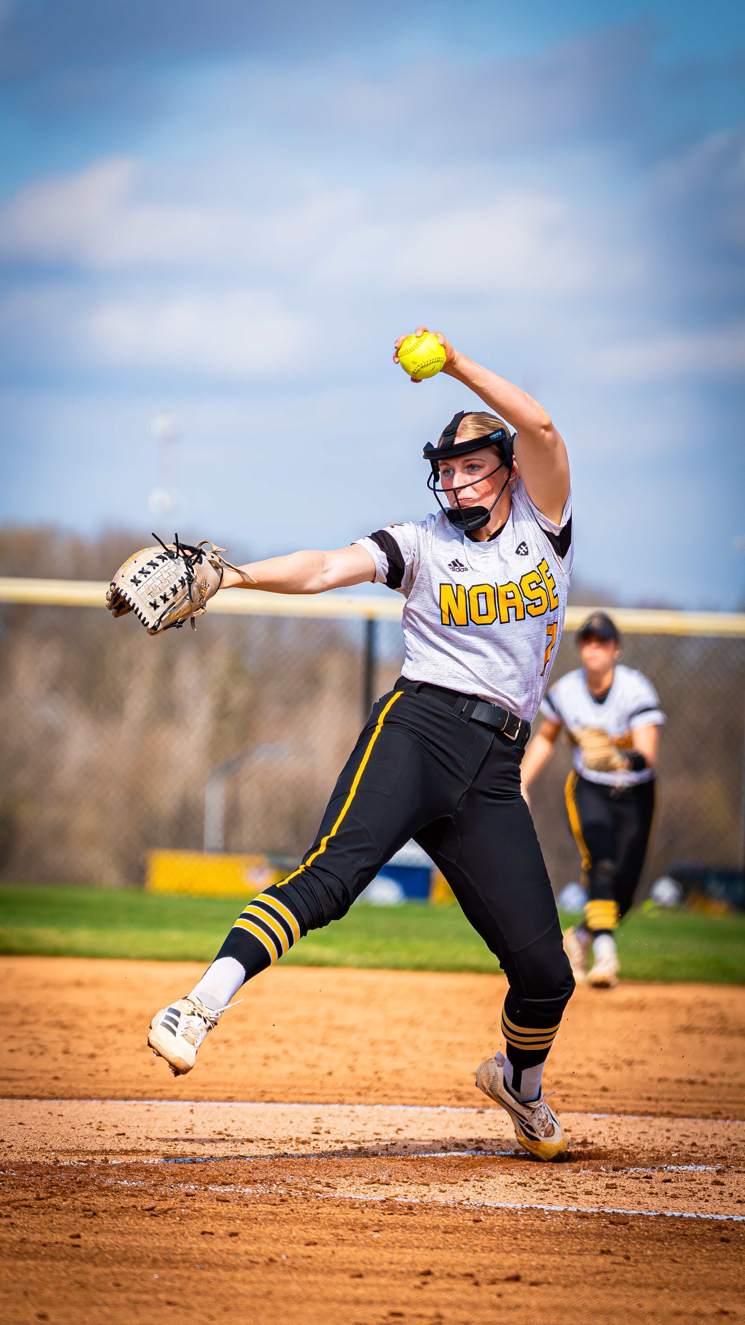 A softball player in black and yellow uniform is pitching the ball on a softball field.