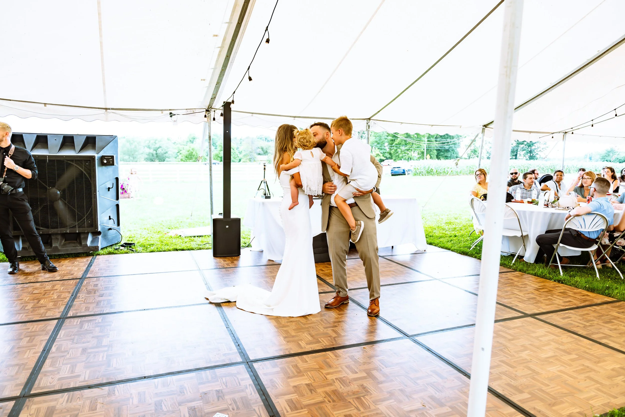 Man and young girl hugging and sharing a kiss at a wedding reception under a white canopy, with guests seated at tables in the background.