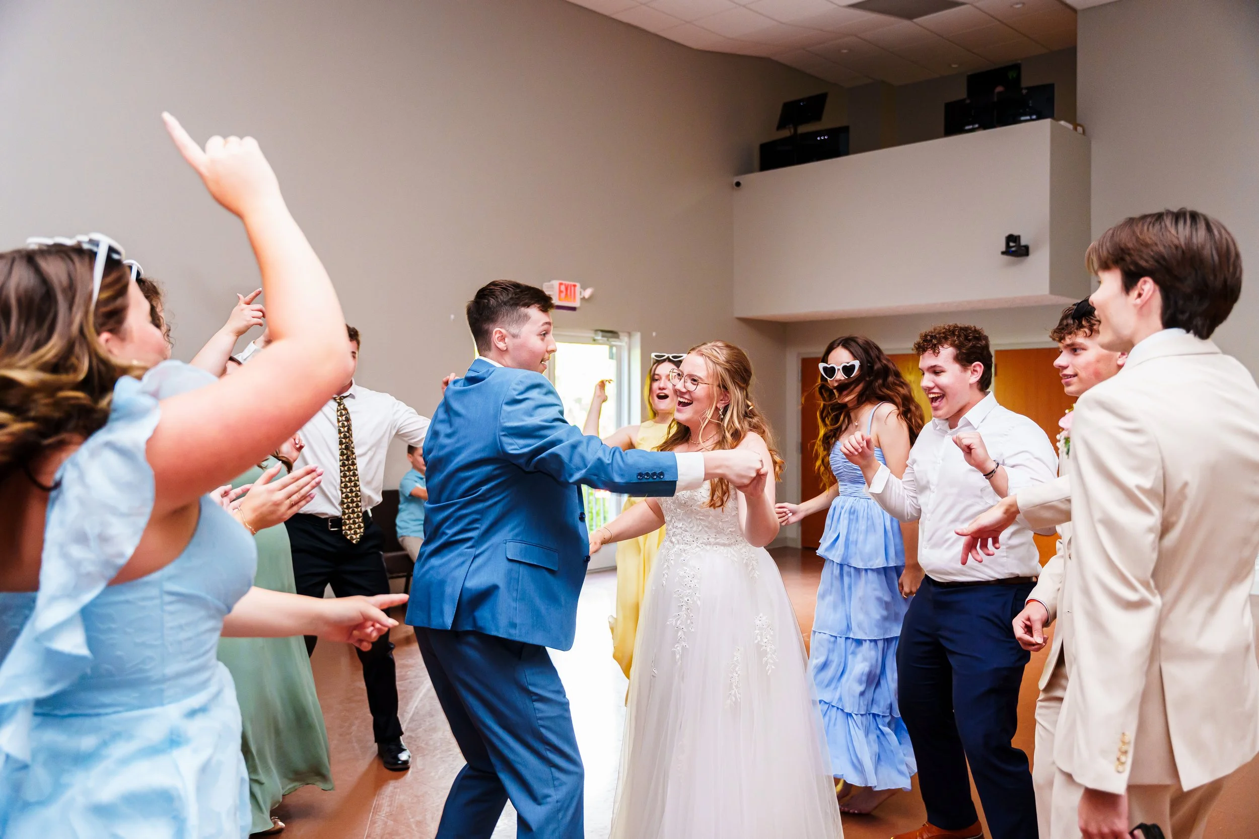 Group of young adults dancing and celebrating at a wedding reception, with some people wearing sunglasses and formal attire.