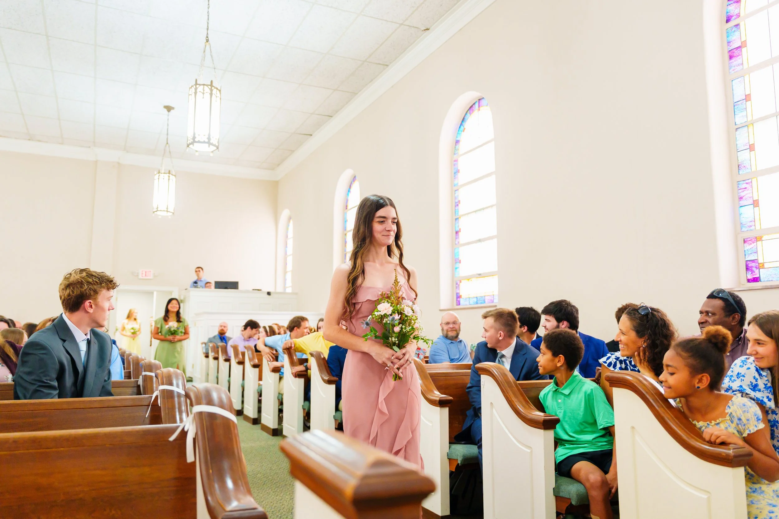 A woman in a pink dress holding a bouquet of flowers walking down the aisle of a church during a wedding ceremony, with guests seated and smiling.