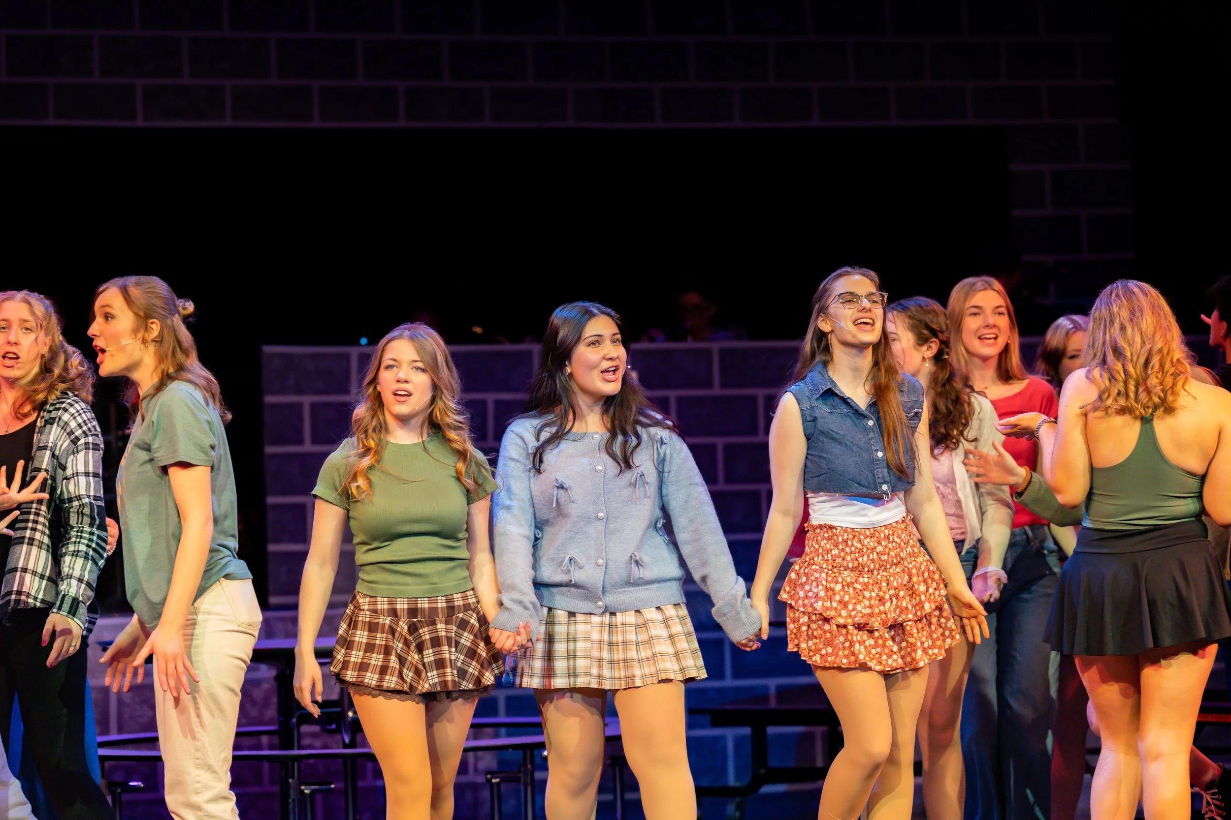 A group of young women standing on stage holding hands, performing in a musical or play, with brick-patterned background.