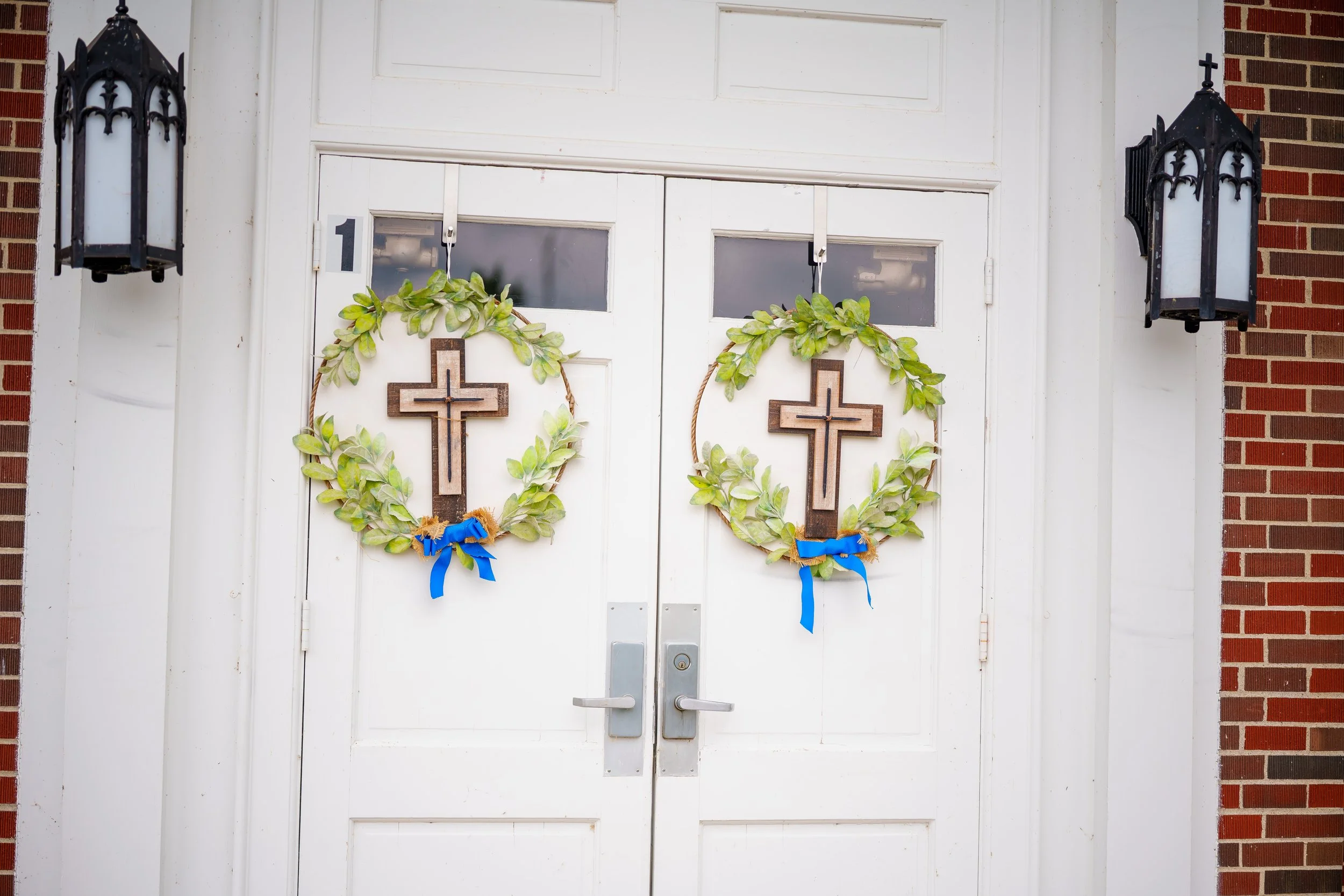 White double doors decorated with two wreaths made of green leaves, each with a wooden cross and a blue ribbon, flanked by black lantern-style exterior lights on a brick wall.