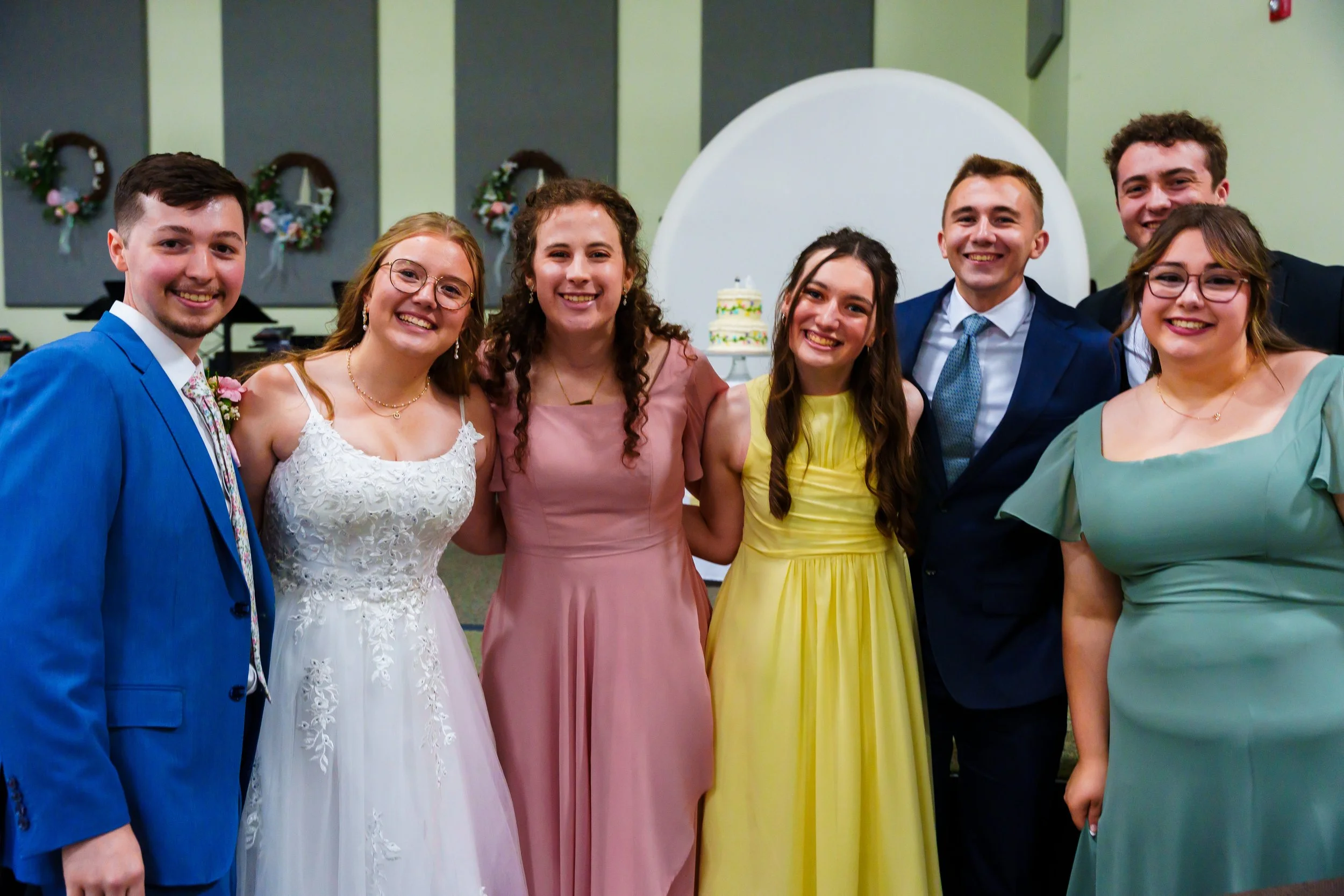 Group of seven young people at a celebration, possibly a wedding, standing together and smiling in an indoor venue decorated with wreaths and a cake in the background.