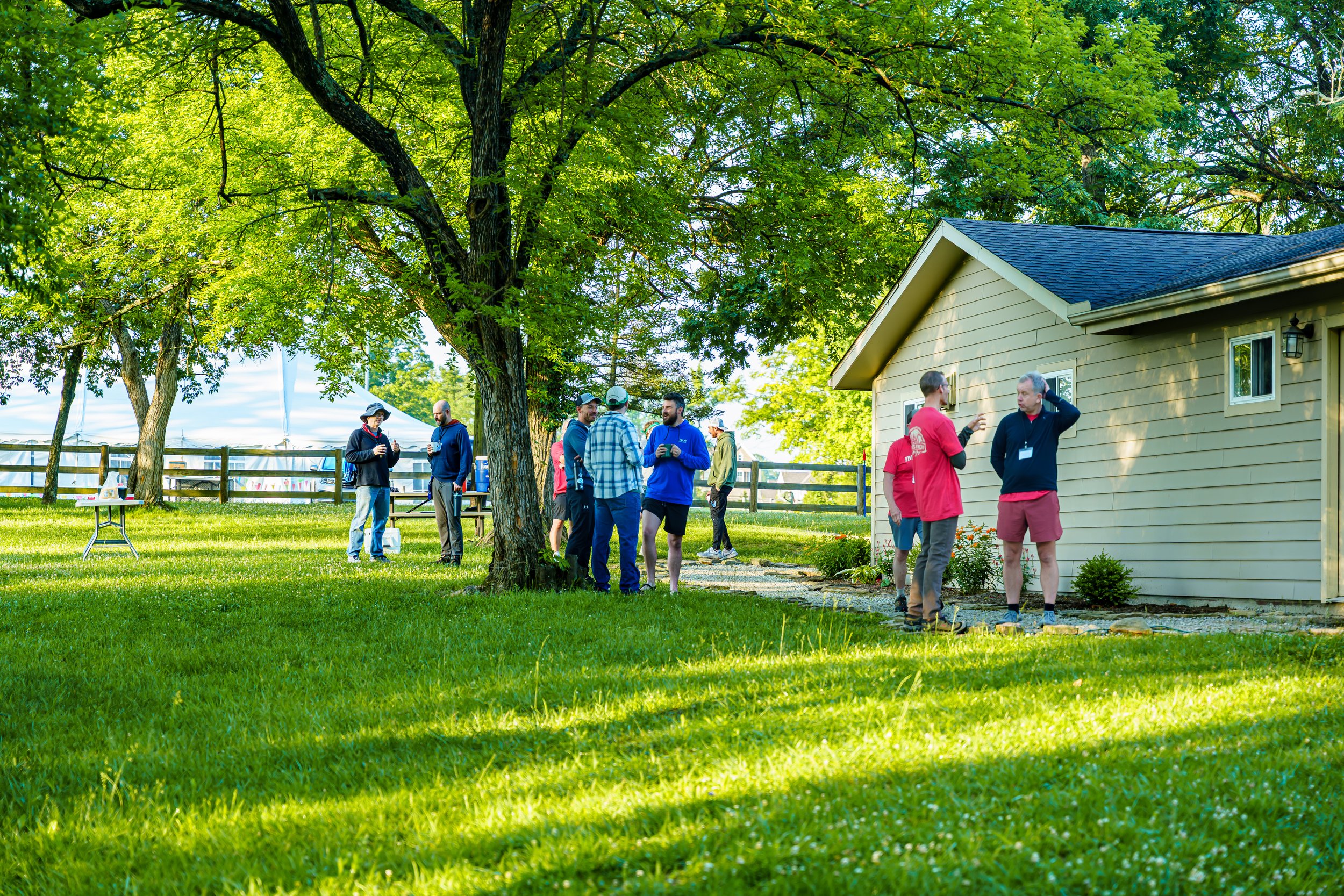 A group of people gathered outdoors near a cream-colored house with a gray roof, under large green trees, on a sunny day.