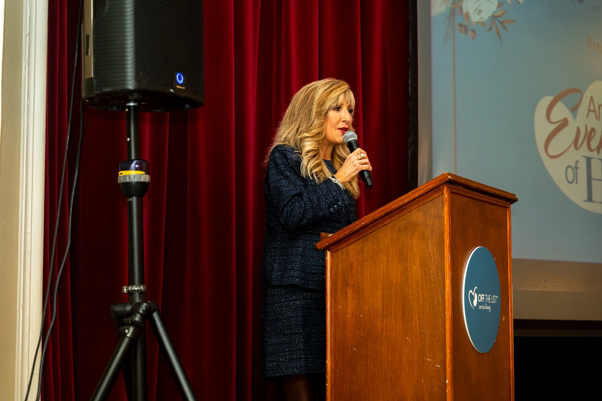 A woman with blonde hair speaking into a microphone at a podium during a presentation with red curtains behind her and a large screen showing a partially visible logo.