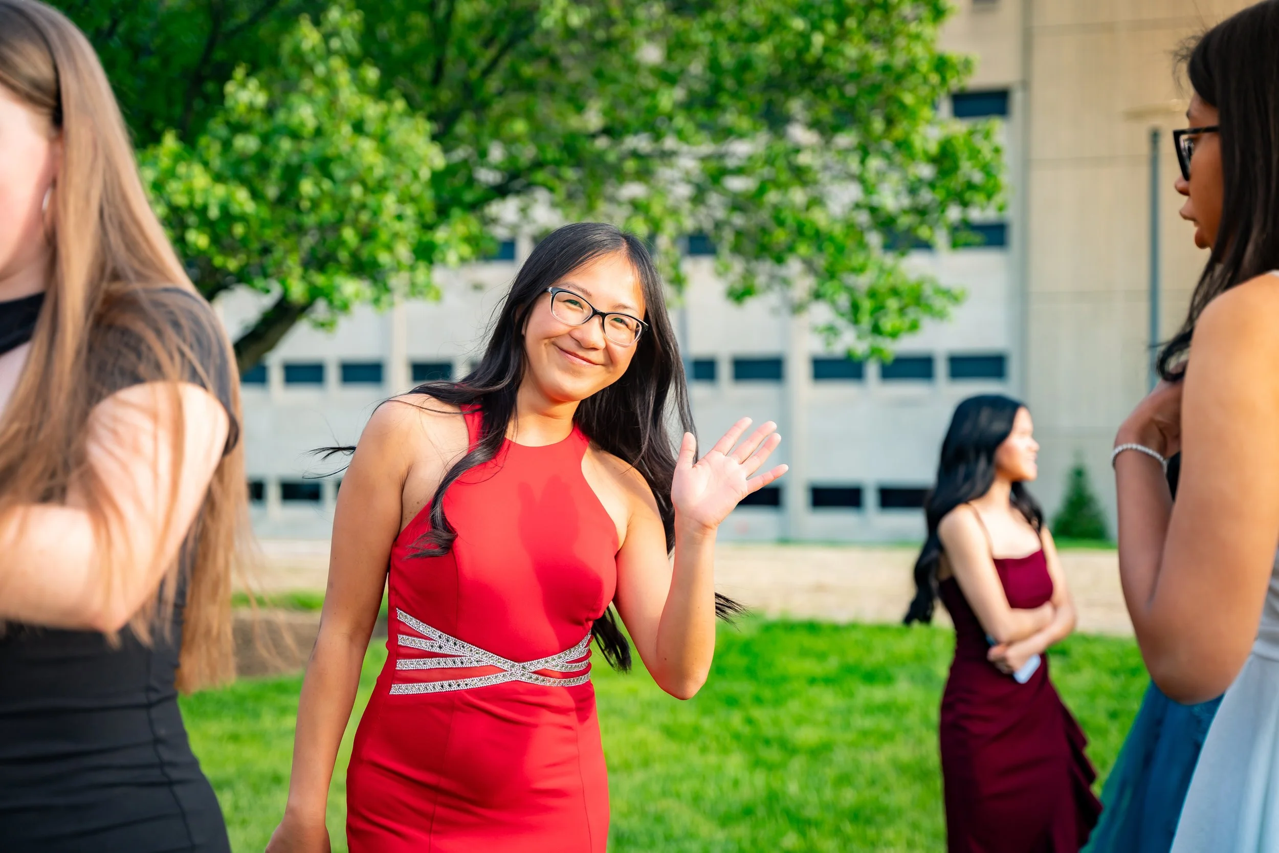 A young woman with glasses and long dark hair smiling and waving to someone, wearing a sleeveless red dress with silver accents, outdoors with green trees and a building in the background.