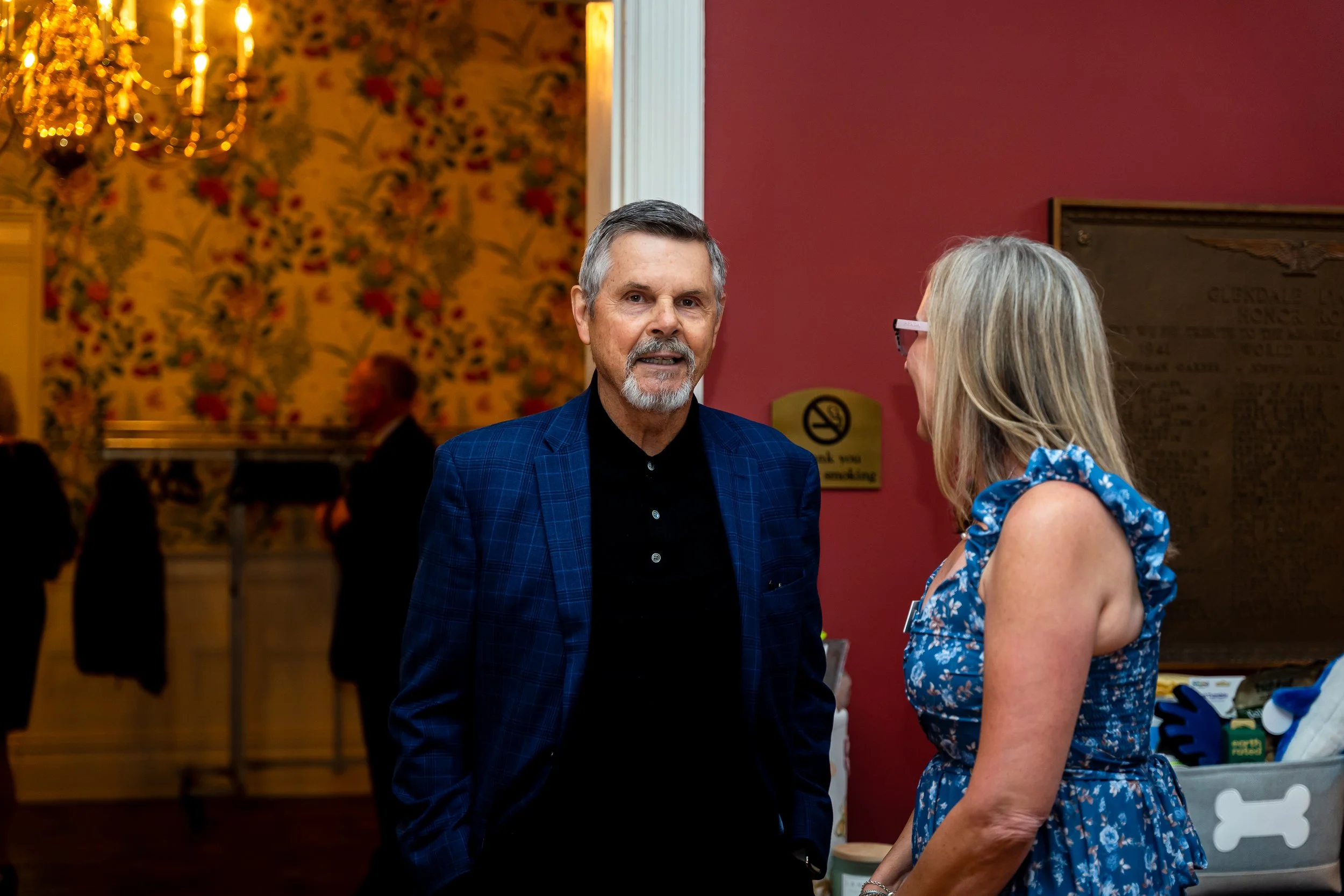 A man with gray hair and a beard wearing a blue checkered blazer talking to a woman with gray hair and glasses, dressed in a blue floral dress, in an indoor setting with a chandelier and floral wallpaper in the background.