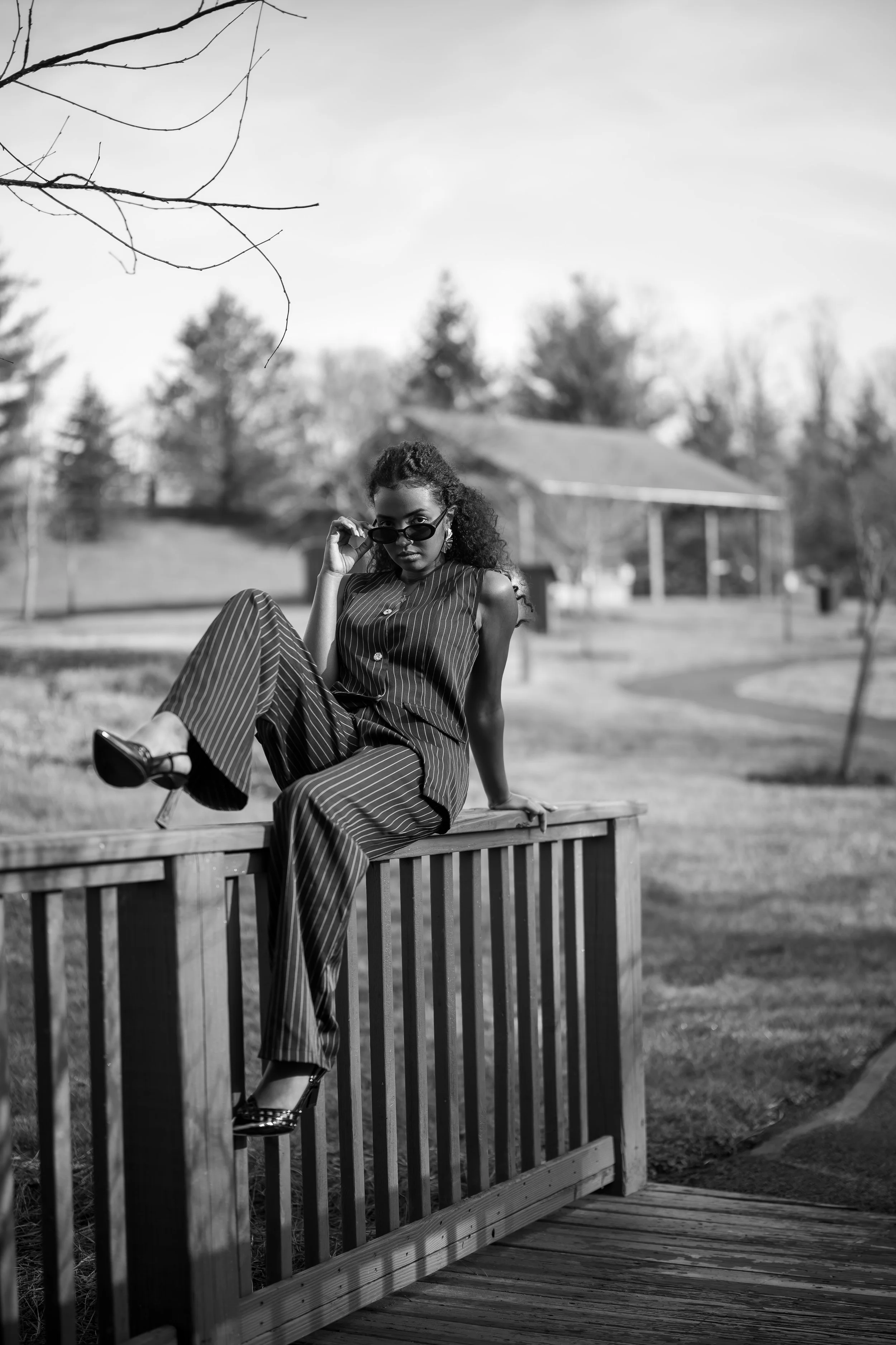 A woman with curly hair wearing sunglasses, a sleeveless striped top, and striped pants, sitting on a wooden railing outdoors with a park and trees in the background.