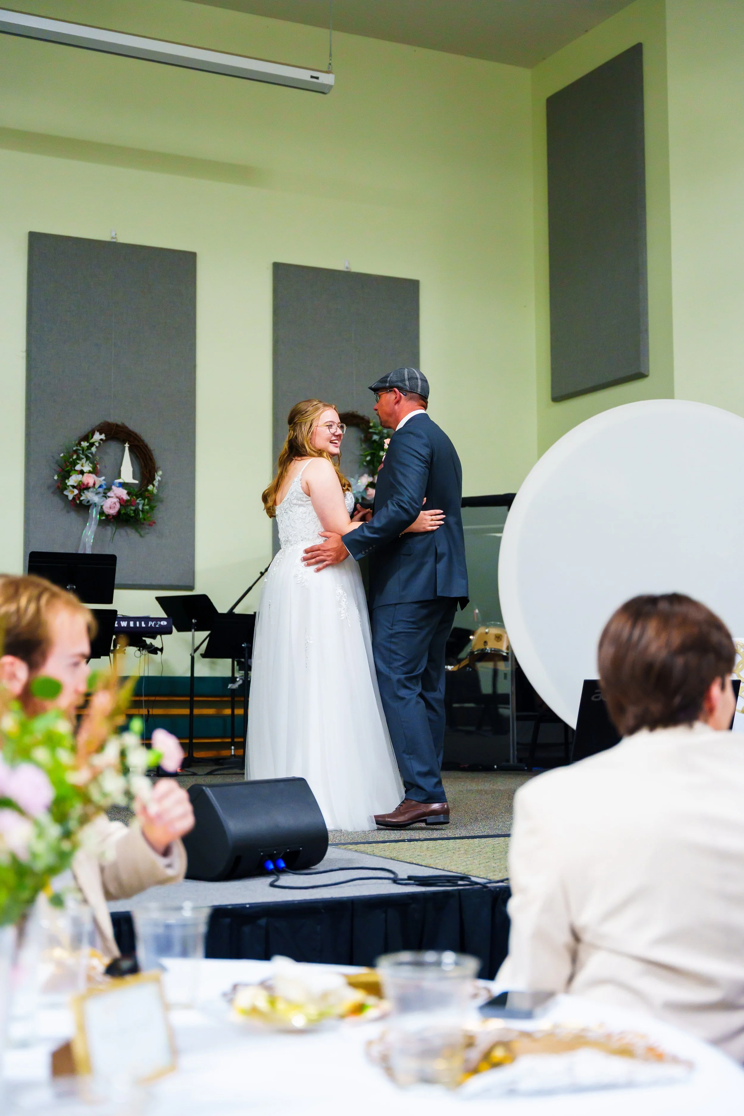 A bride and groom are dancing during their wedding ceremony inside a decorated hall. The bride wears a white gown and the groom wears a dark suit with a flat cap. Guests are seated at decorated tables, and there are musical instruments and speakers i