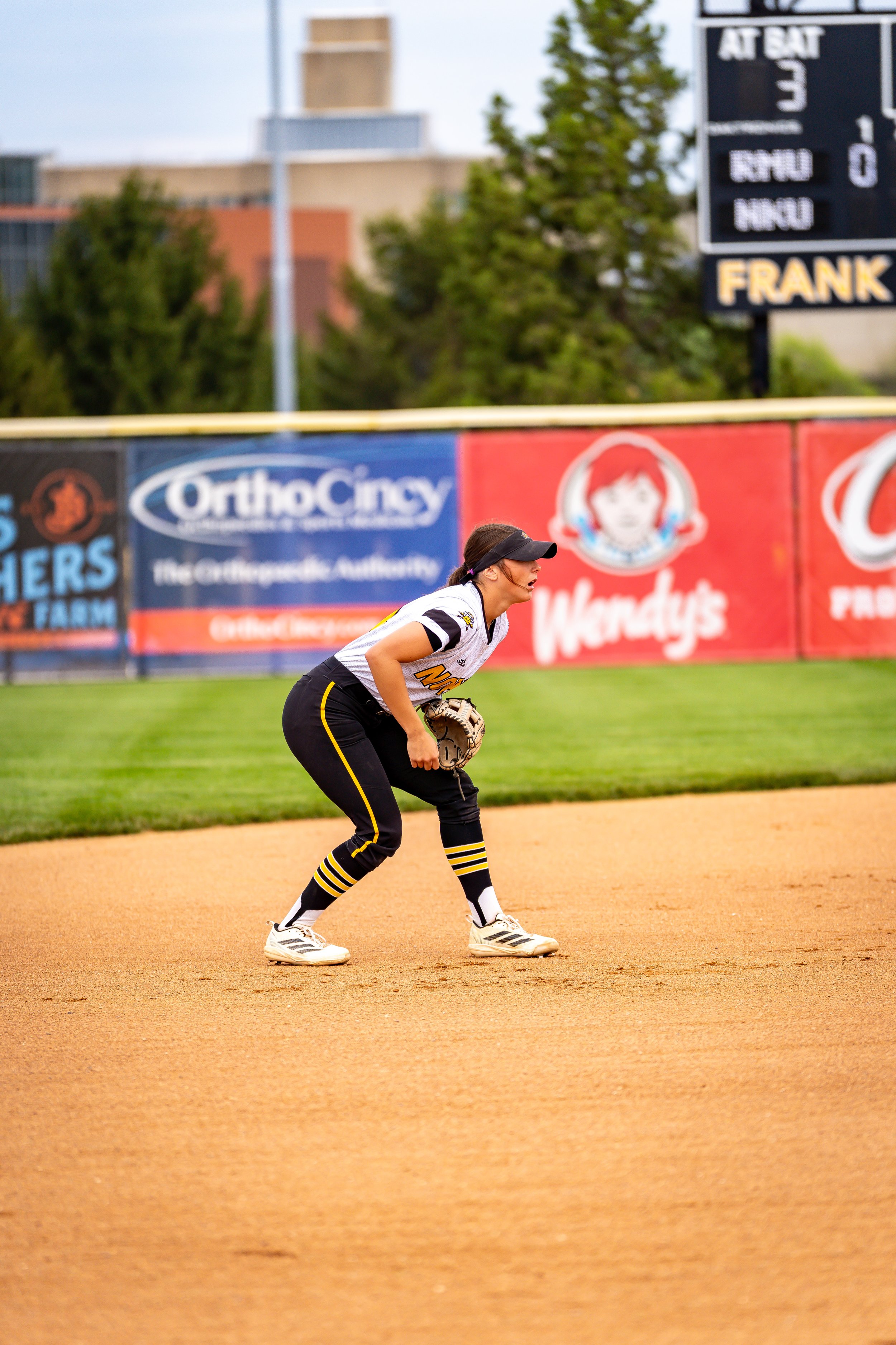 A female softball player is in a ready stance on the field, wearing a black and yellow uniform with a black visor and white cleats, against a background of advertisement banners, trees, and a scoreboard.