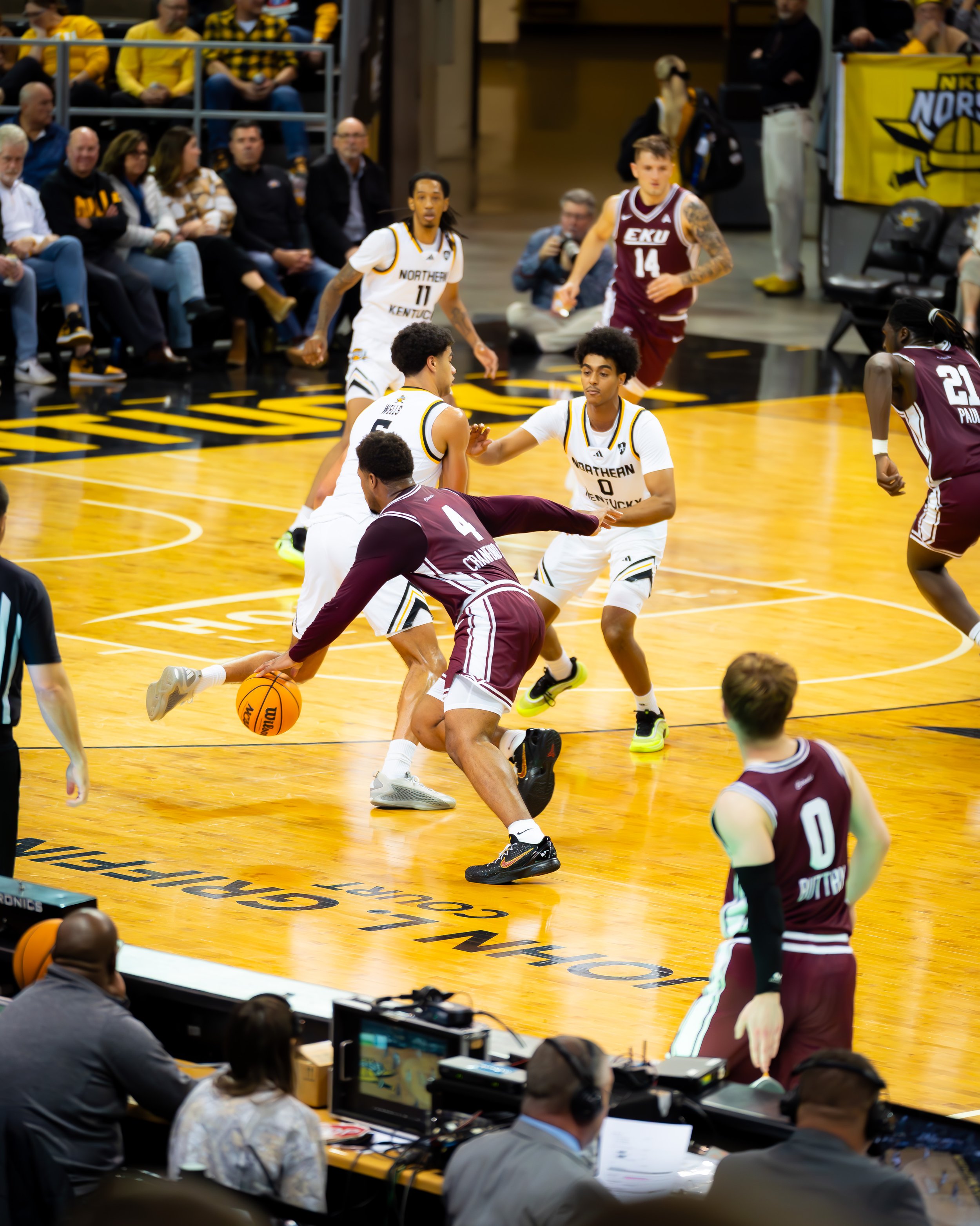 College basketball game with players from Northern Kentucky and EKU on the court. A player from EKU is dribbling the ball while defenders from Northern Kentucky attempt to block him. Spectators are seated in the background, and officials and broadcas