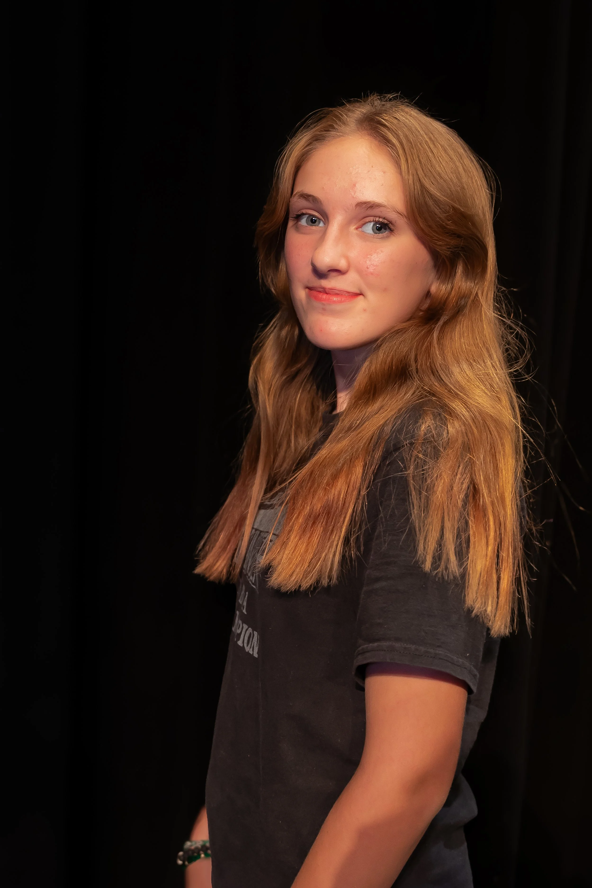 Close-up of a teenage girl with long, wavy, reddish-brown hair, blue eyes, and light skin, smiling slightly against a black background.