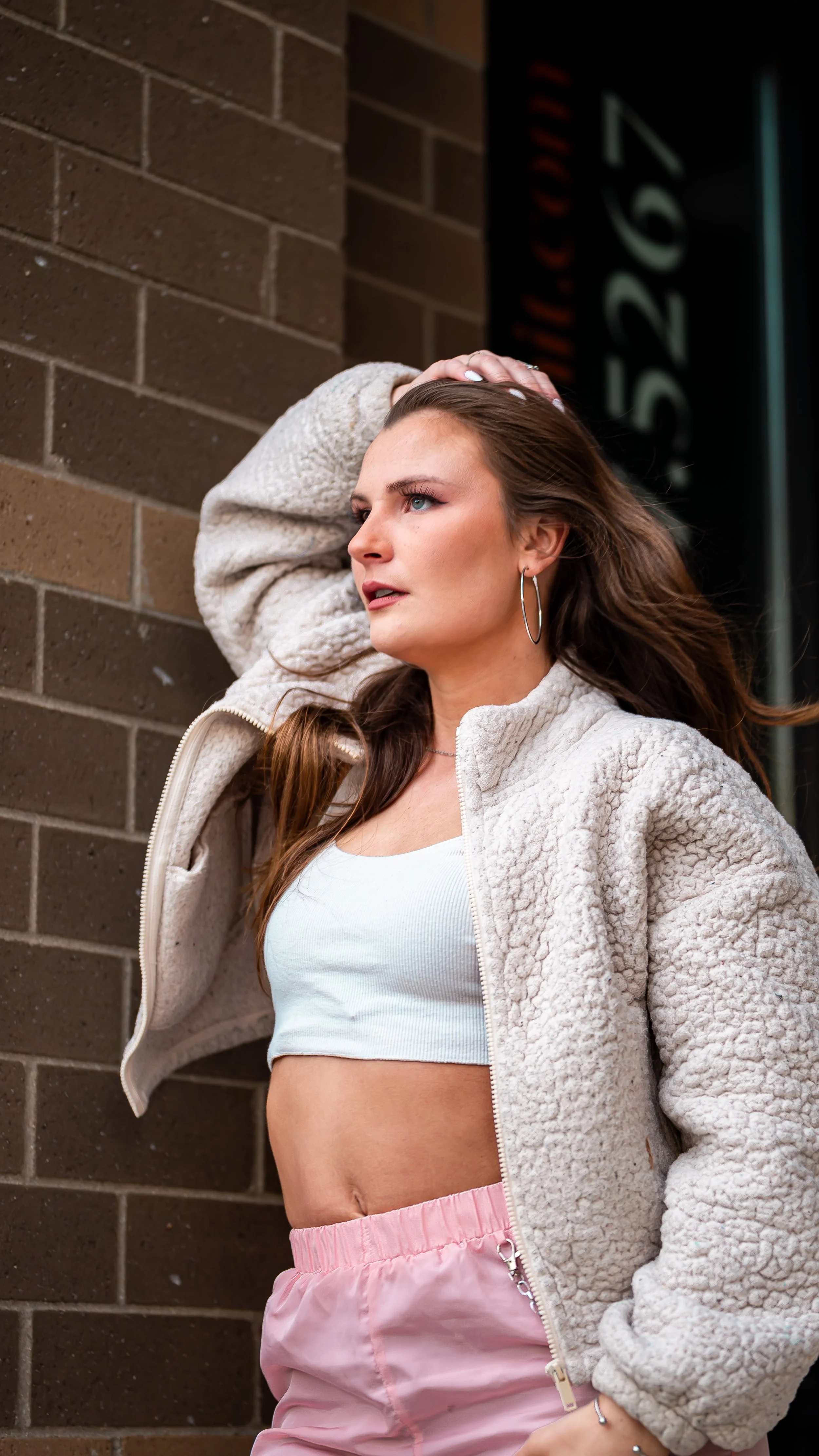 A young woman with long brown hair and blue eyes is posing against a brick wall, wearing a beige sherpa jacket, a white crop top, pink shorts, and hoop earrings.