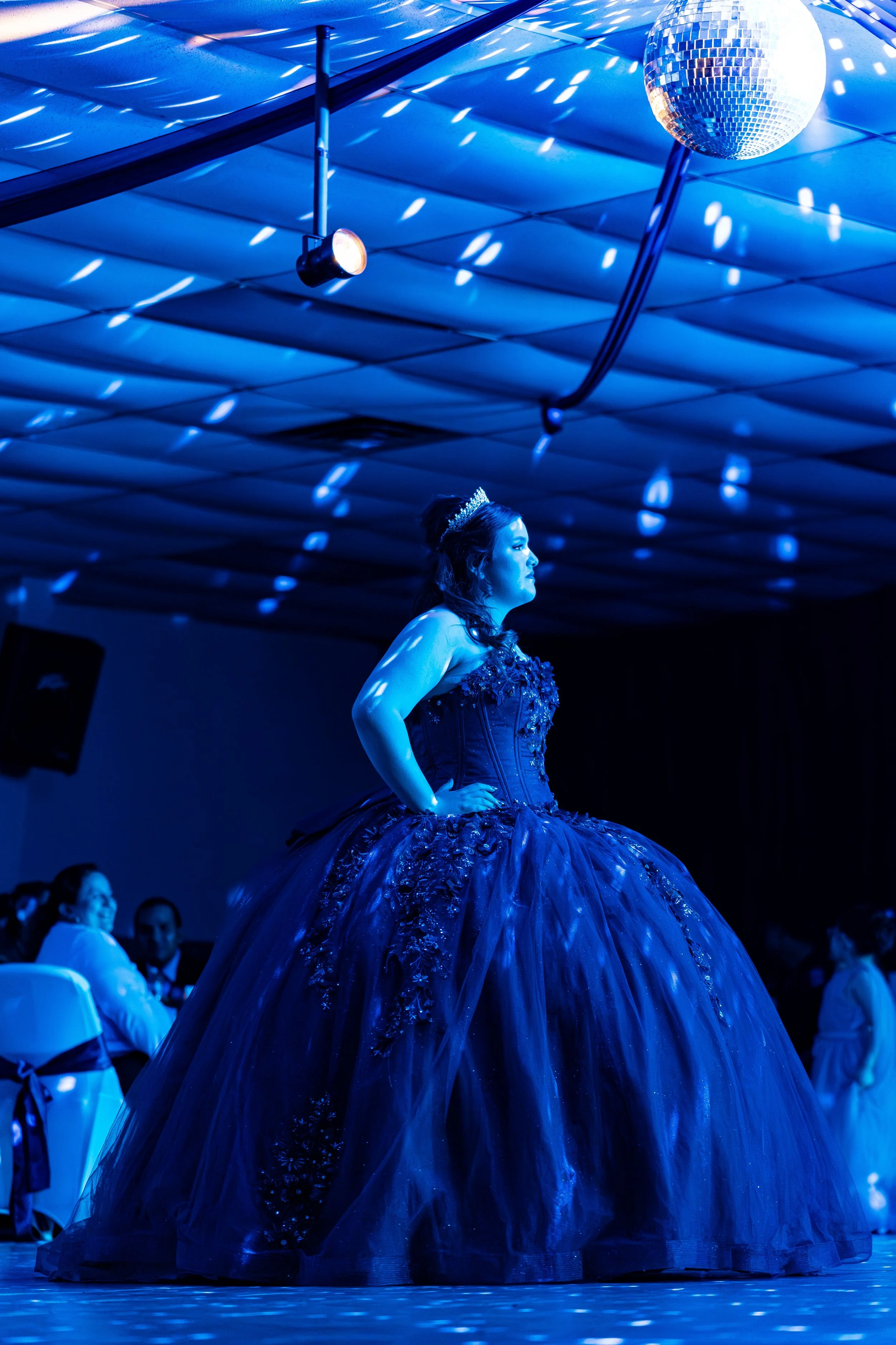 A woman in an elaborate, dark blue ball gown with floral embellishments, standing with her hands on her hips under blue lighting and a disco ball at the ceiling.