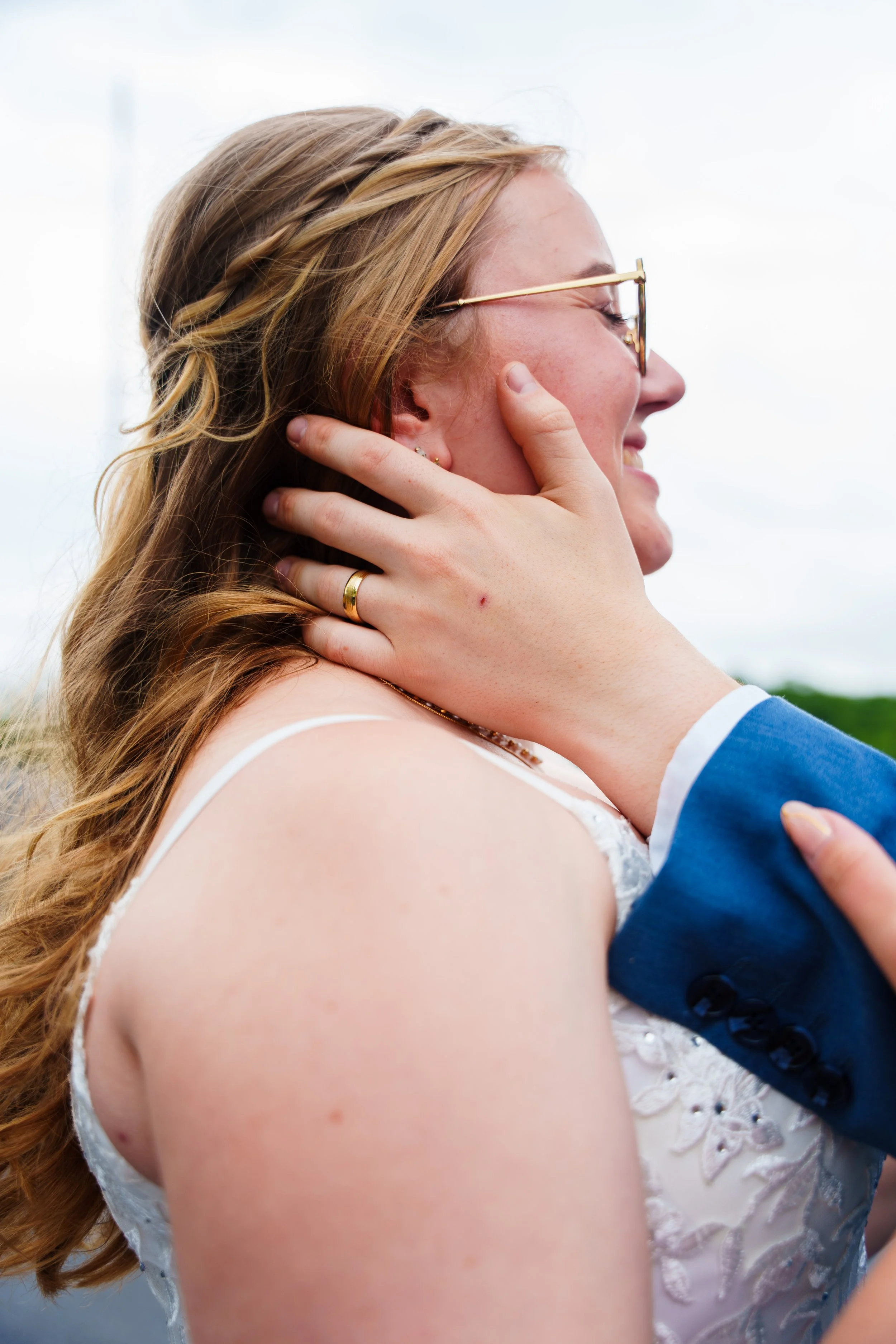 A woman and man embrace outdoors, close-up of faces, woman with glasses and lace dress, man in blue suit, sky in background.
