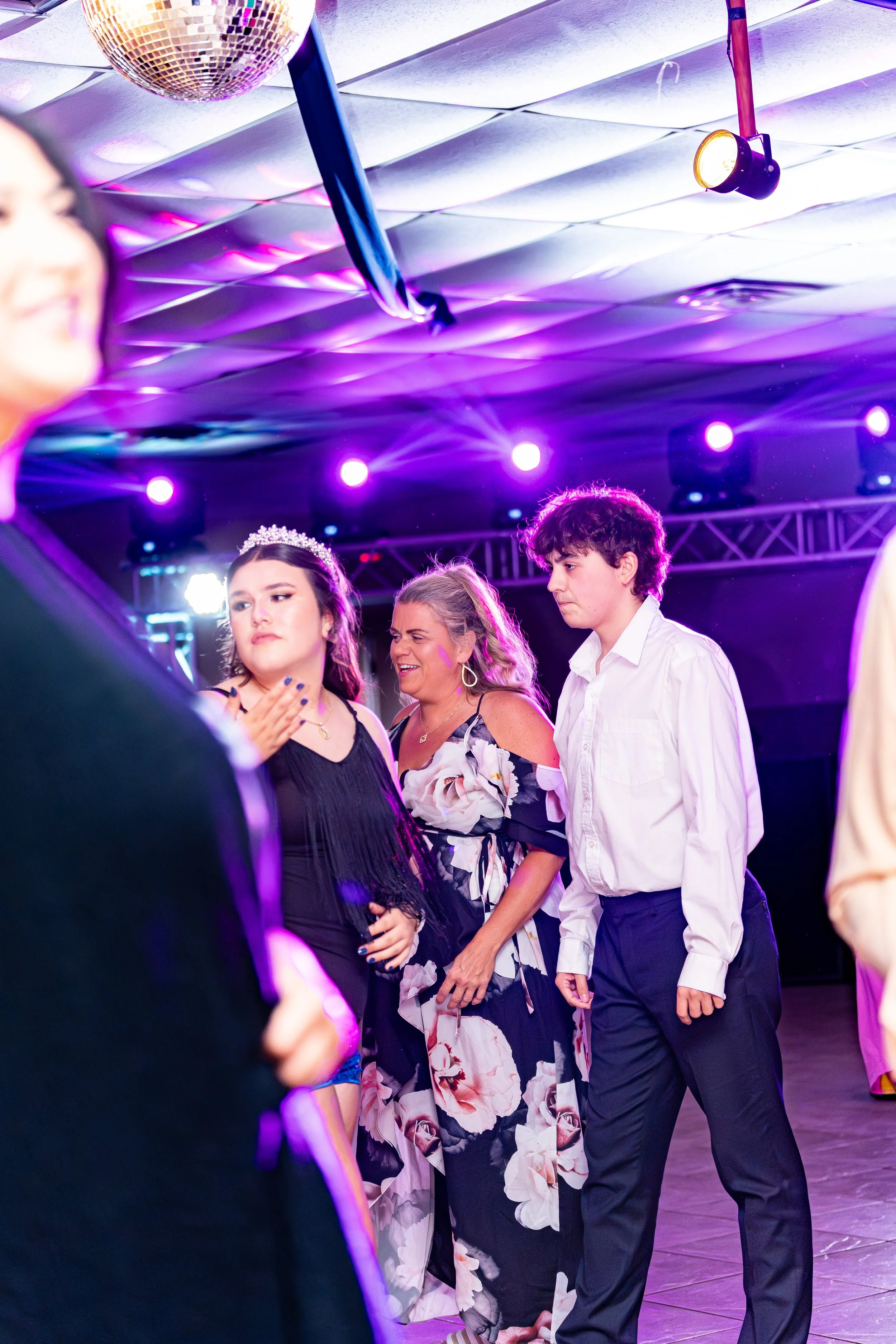 People dancing at a party or dance event with colorful lighting, including a woman wearing a tiara and floral dress, and a young man in a white shirt and dark pants.