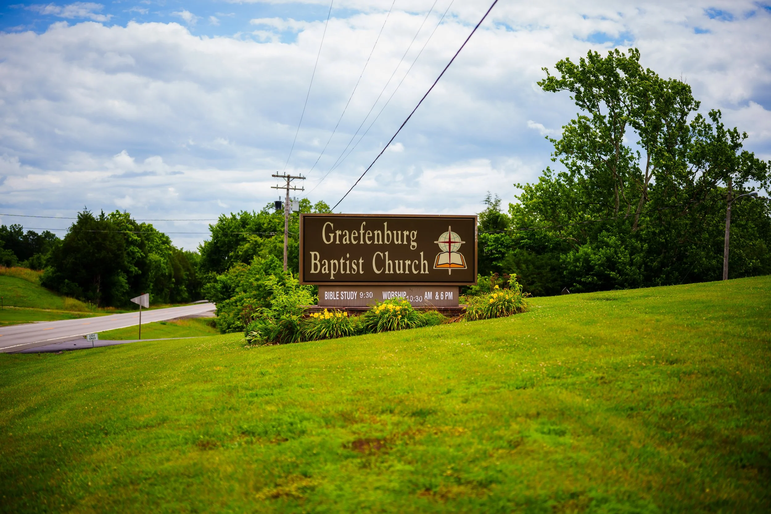 Sign for Graefenburg Baptist Church with service times and a religious symbol, set on a grassy area with trees and a cloudy sky in the background.