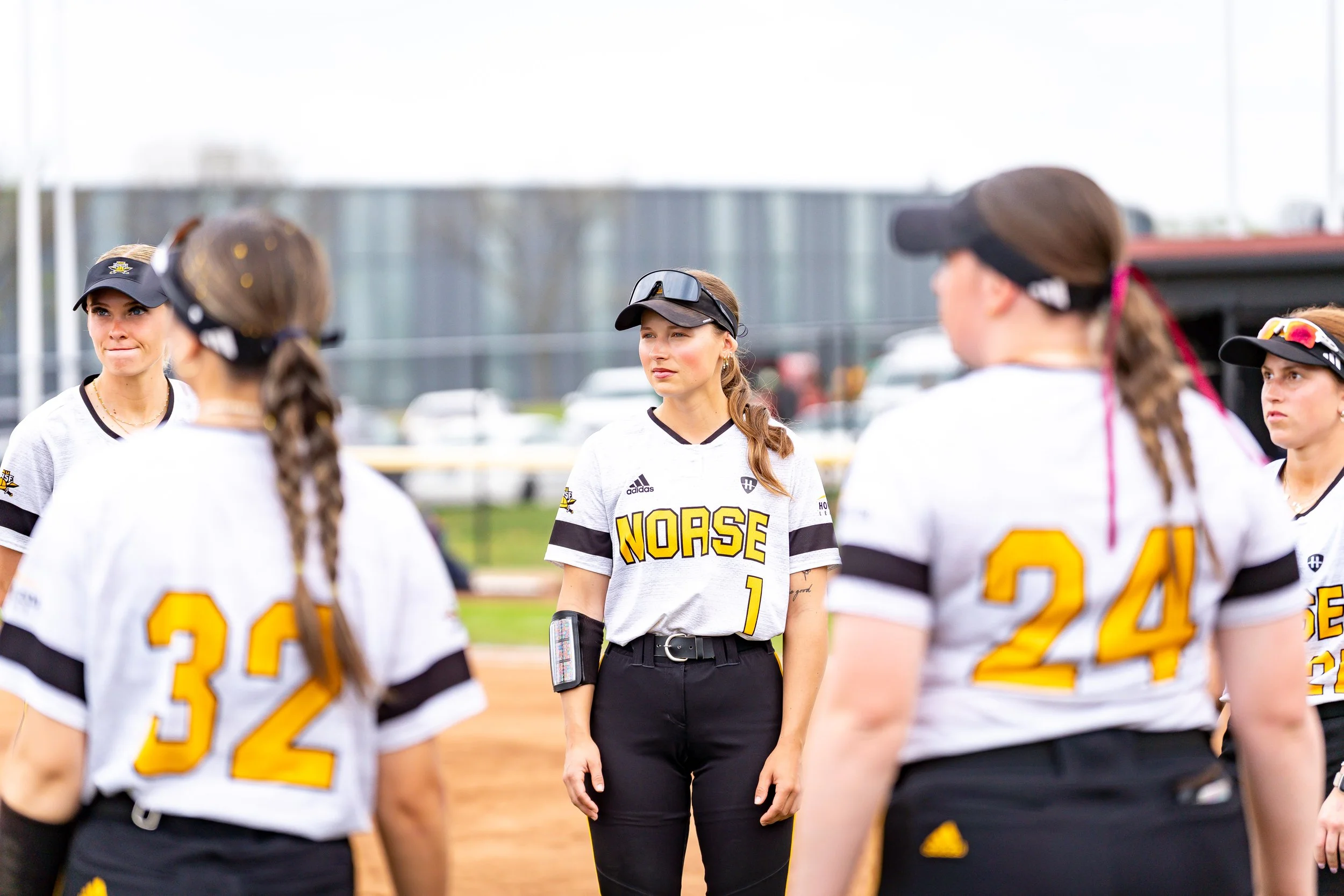 A group of female softball players in white jerseys with yellow and black lettering and numbers, gathered around a woman in black pants and a black visor, likely a coach, on a softball field.