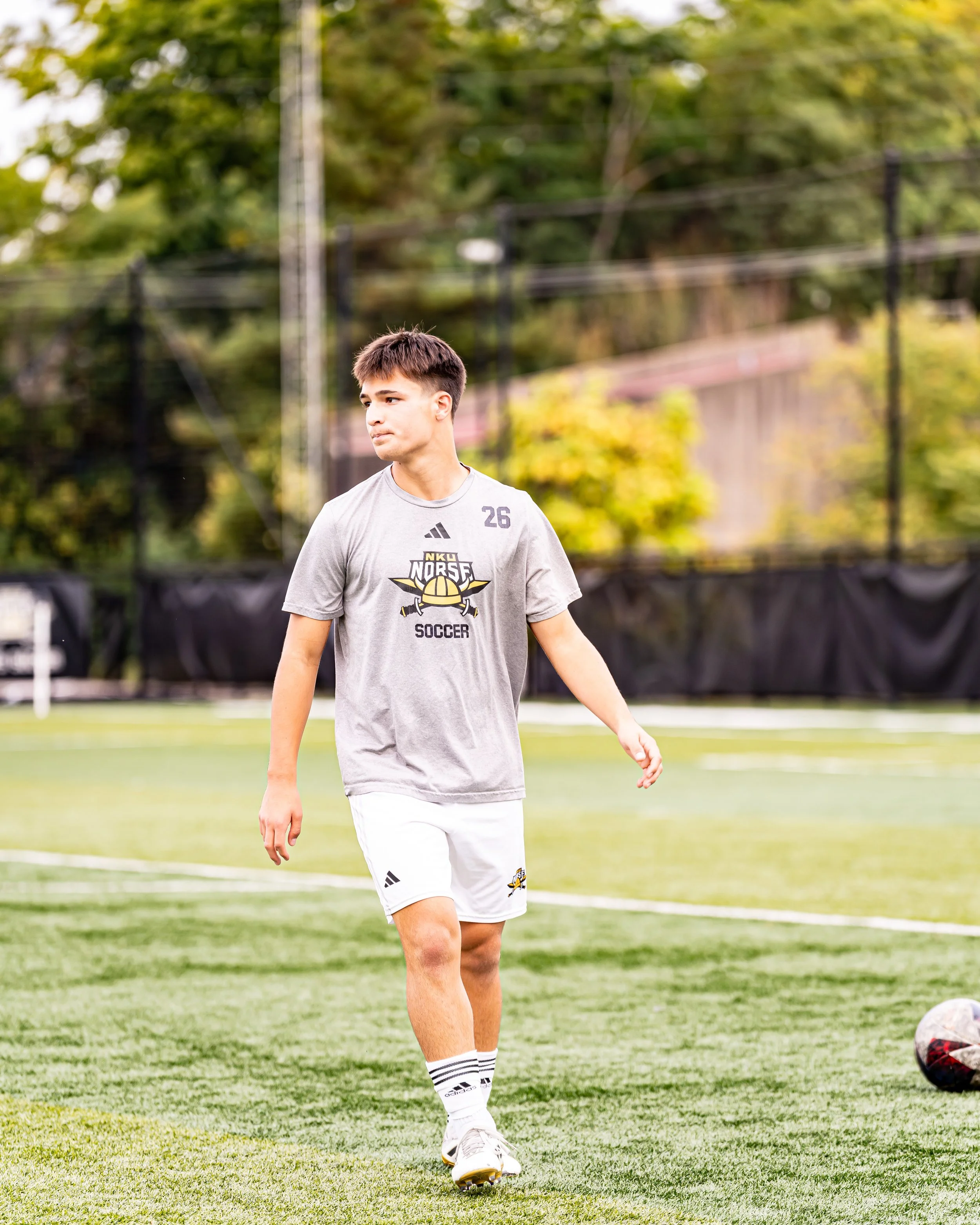 A young male soccer player walking on a soccer field during practice or game, wearing a grey t-shirt, white shorts, and white Adidas socks, with a soccer ball on the ground nearby.